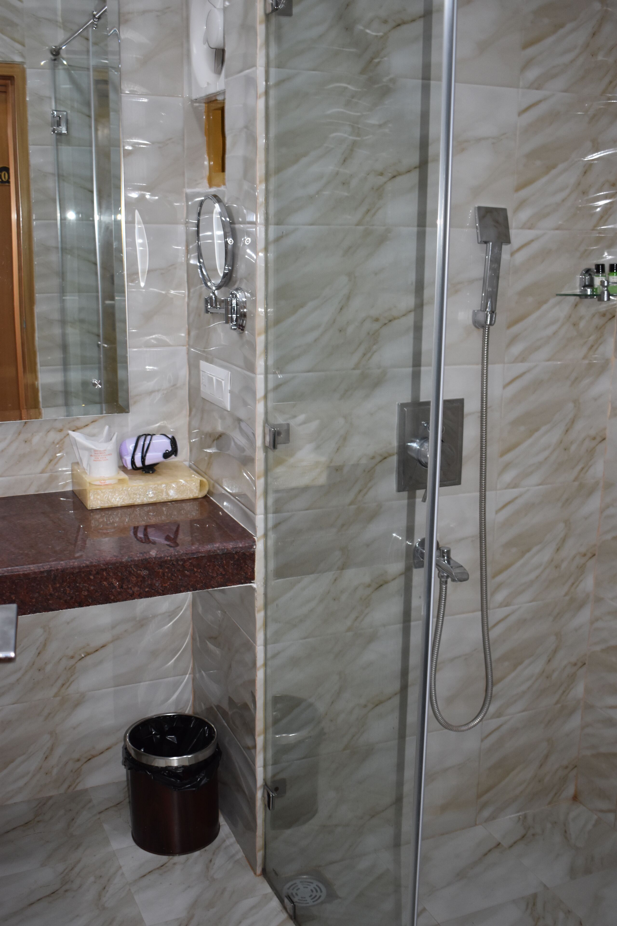 Un baño con pared de mármol, cabina de ducha de vidrio, lavamanos con espejo y un cubo de basura.