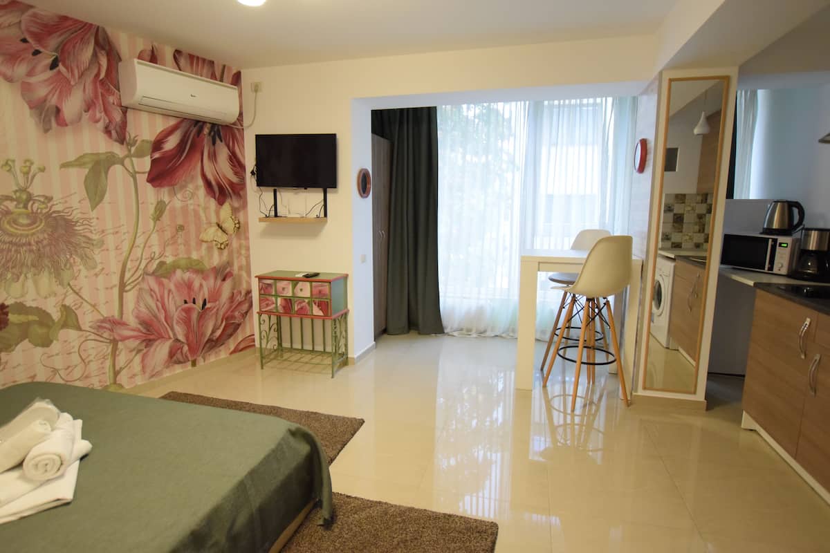 A hotel room with a bed, a small table, a chair, a TV, a kitchen area, and a bathroom visible through an open door.