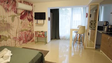 A hotel room with a bed, a small table, a chair, a TV, a kitchen area, and a bathroom visible through an open door.