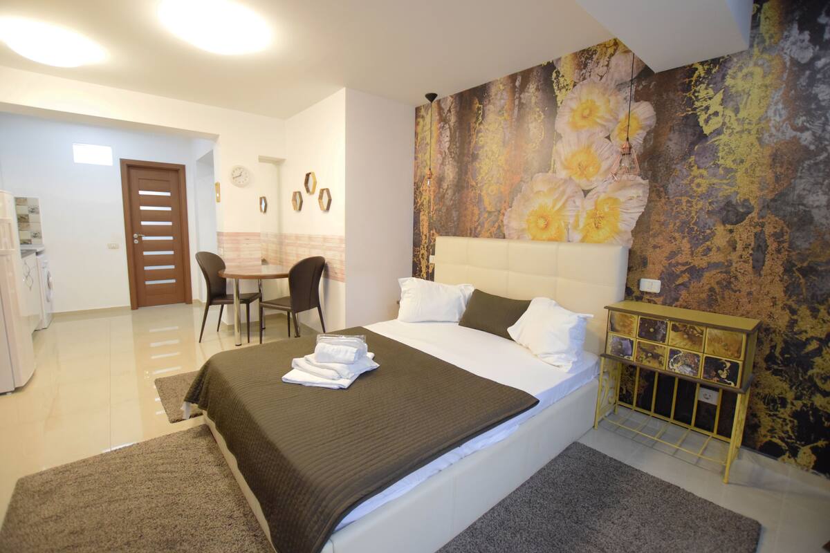 A bedroom with a large bed, a bedside table, a chair, a desk, and a wall with a floral mural.