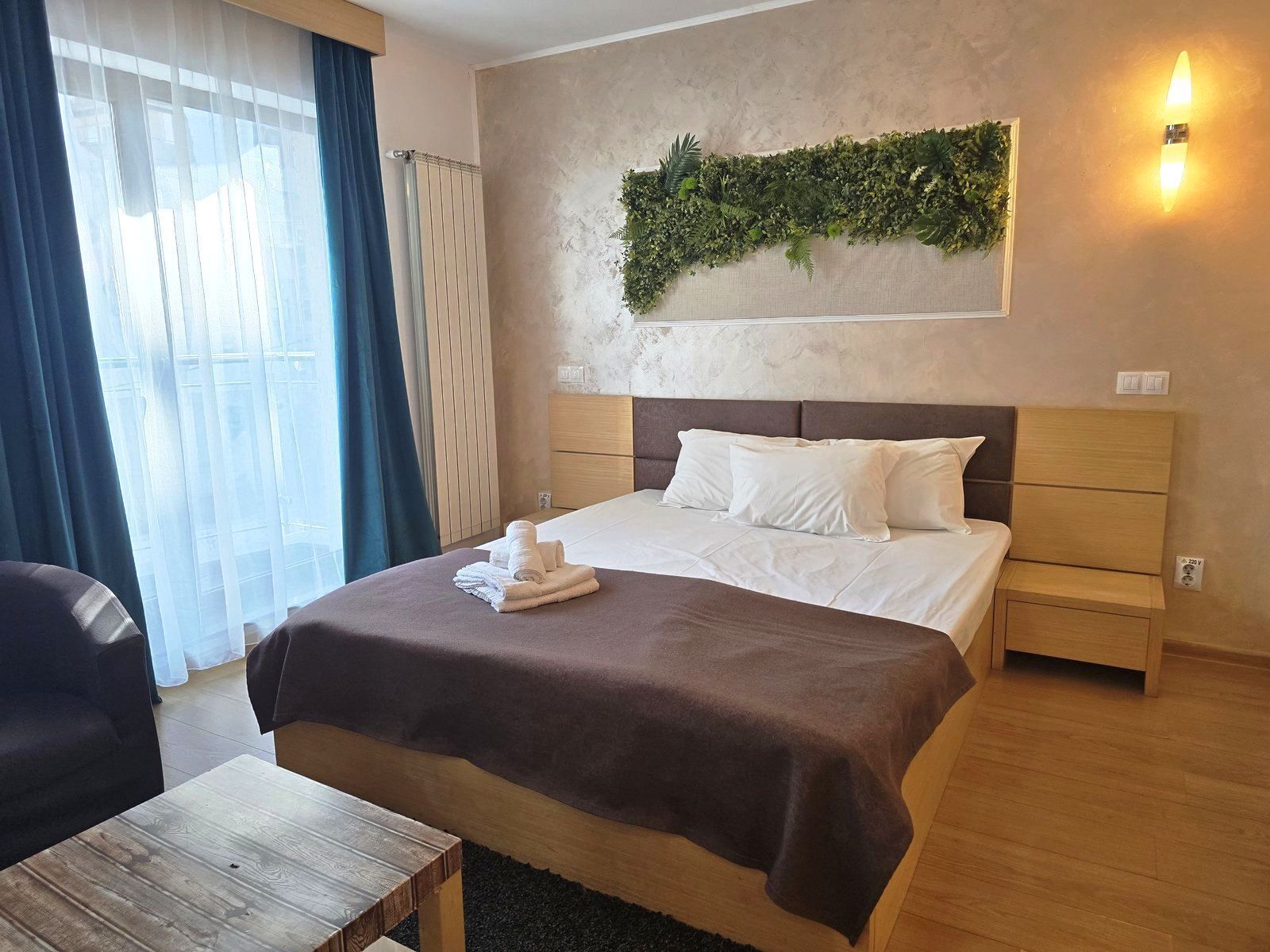 A hotel room with a bed, bedside tables, a small wooden table, a window with curtains, and a wall-mounted plant.