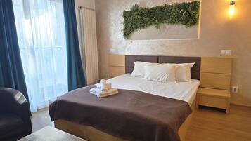 Quarto de hotel com cama, mesas de cabeceira, uma pequena mesa de madeira, janela com cortinas e uma planta fixada na parede.