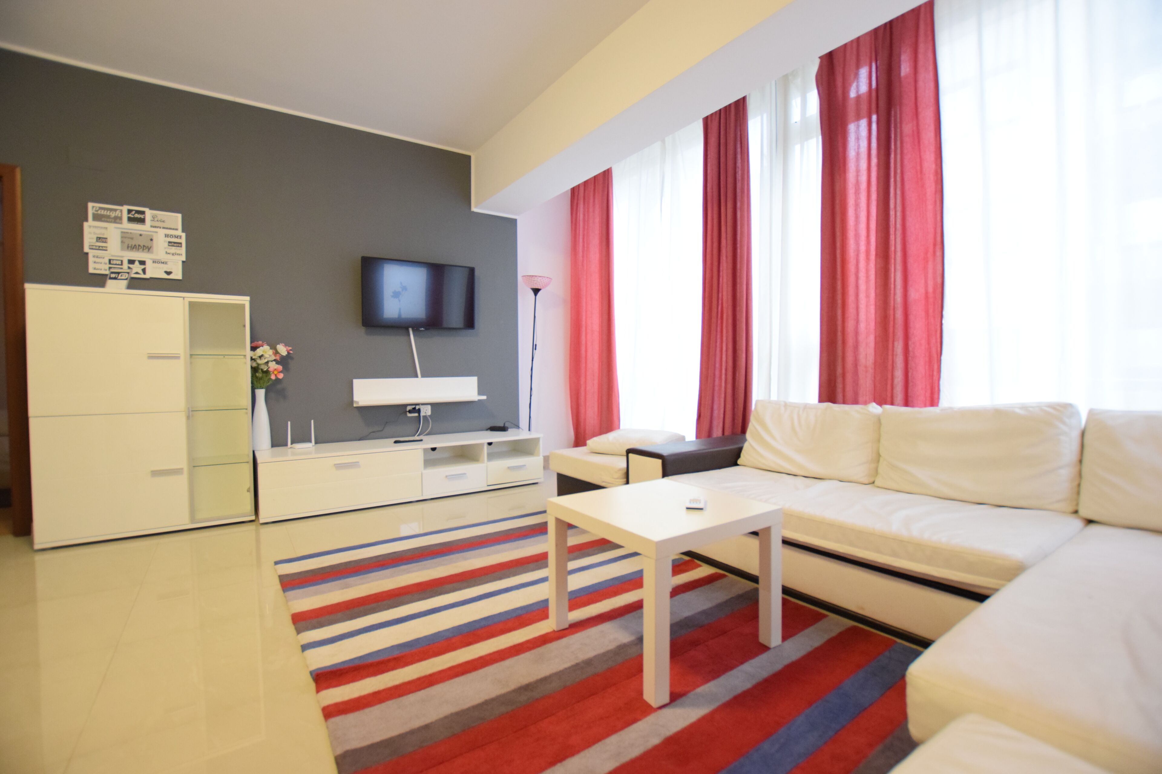A modern living room with a sofa, a coffee table, a TV mounted on the wall, and a large window with red curtains.