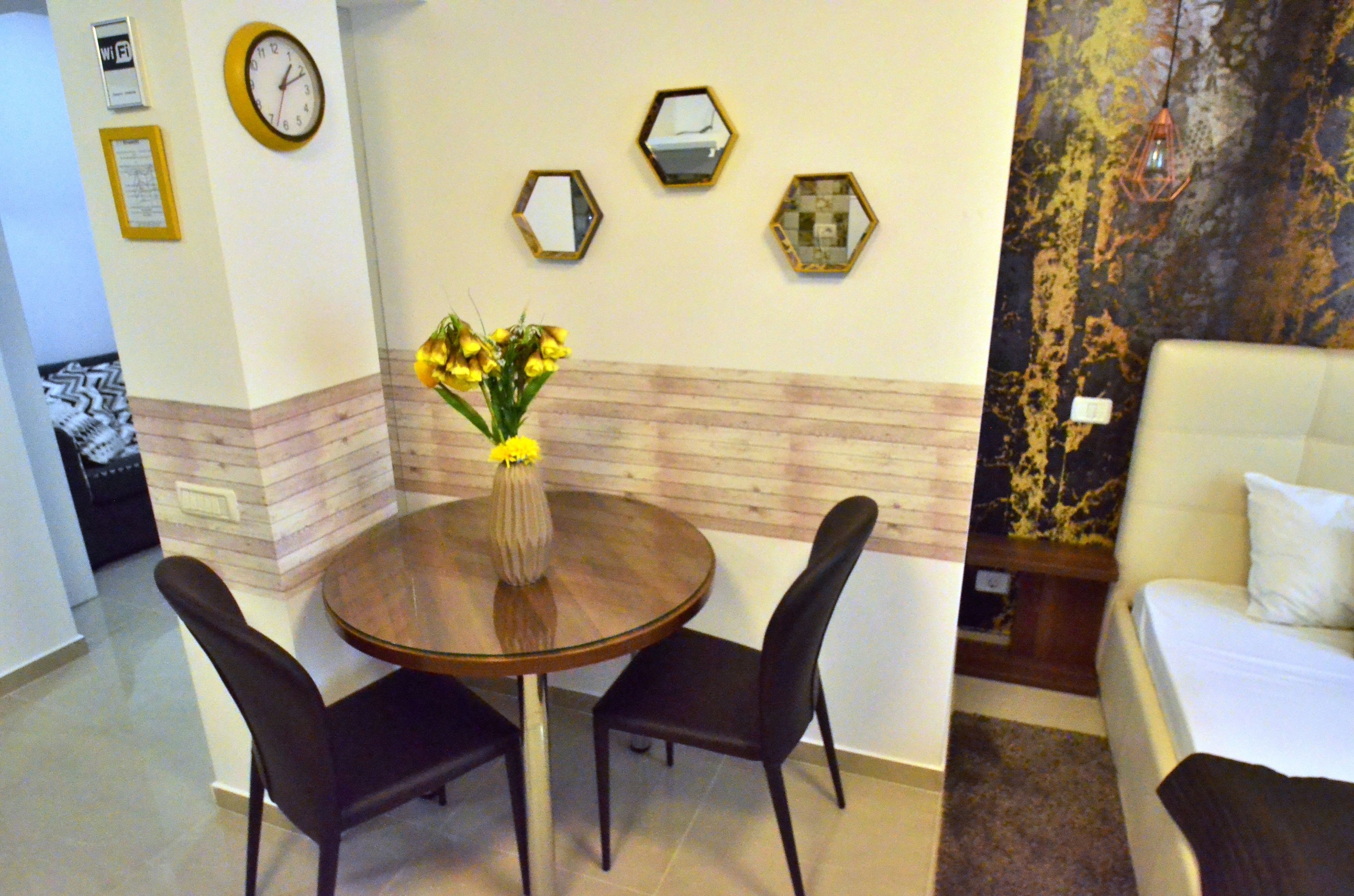 A hotel room with a round dining table, two chairs, a vase with flowers, a bed, and a wall with hexagonal mirrors.