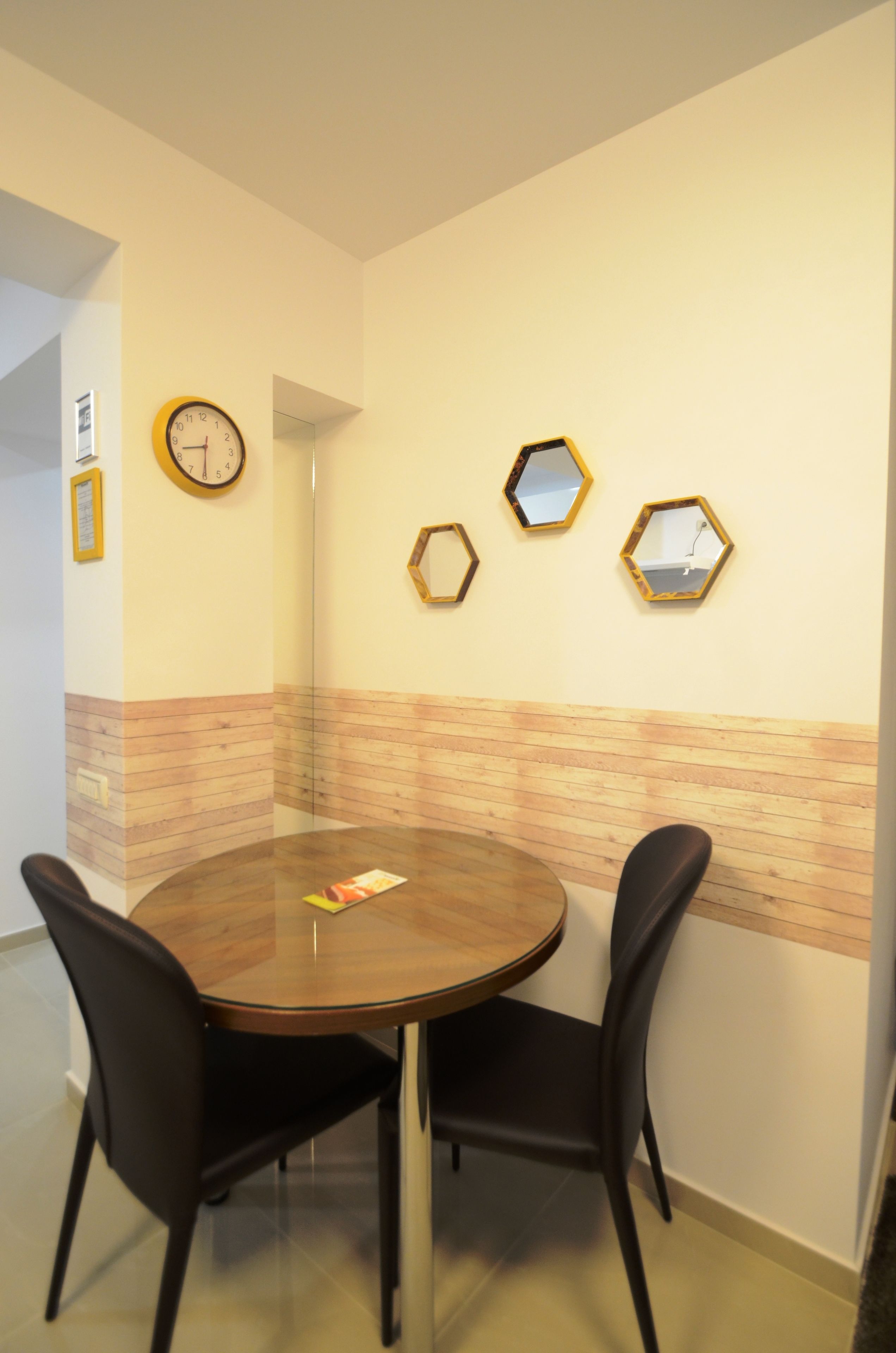 A dining area with a round table, two black chairs, a clock, and three hexagonal mirrors on the wall.