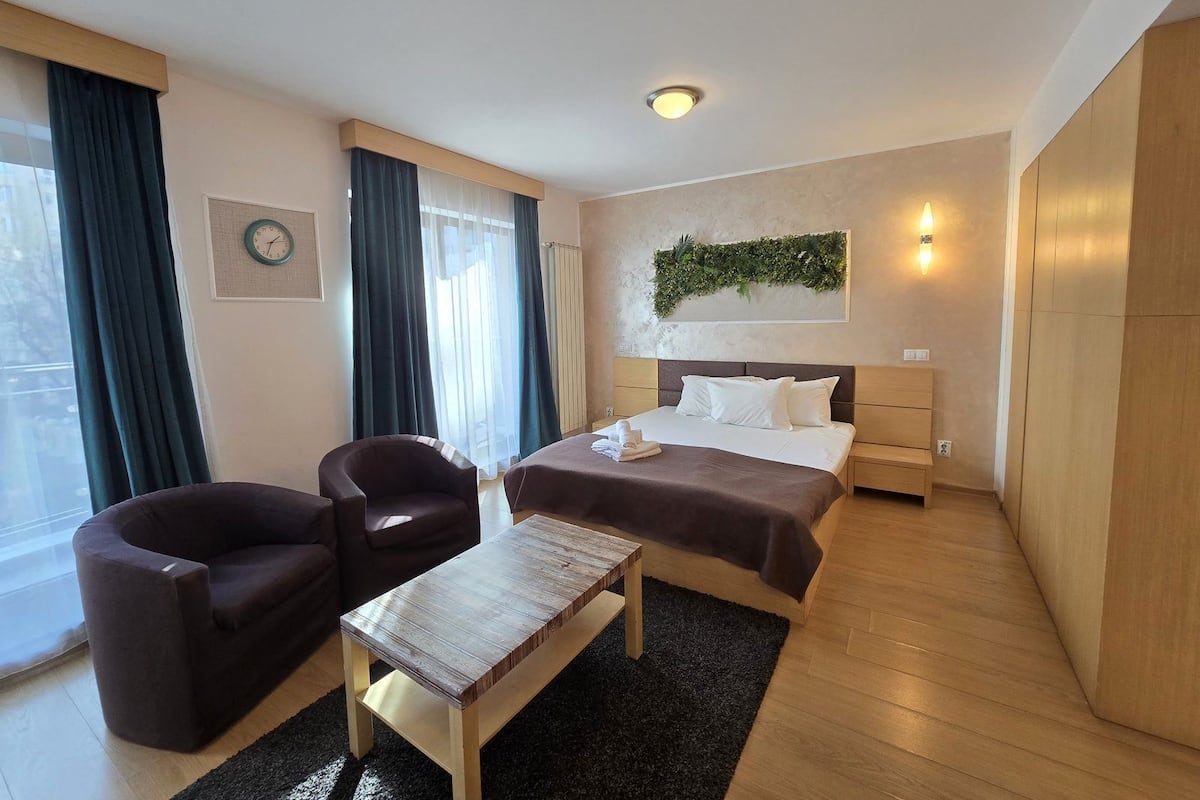 A modern hotel room with a bed, two armchairs, a small wooden table, a clock on the wall, and a decorative plant.