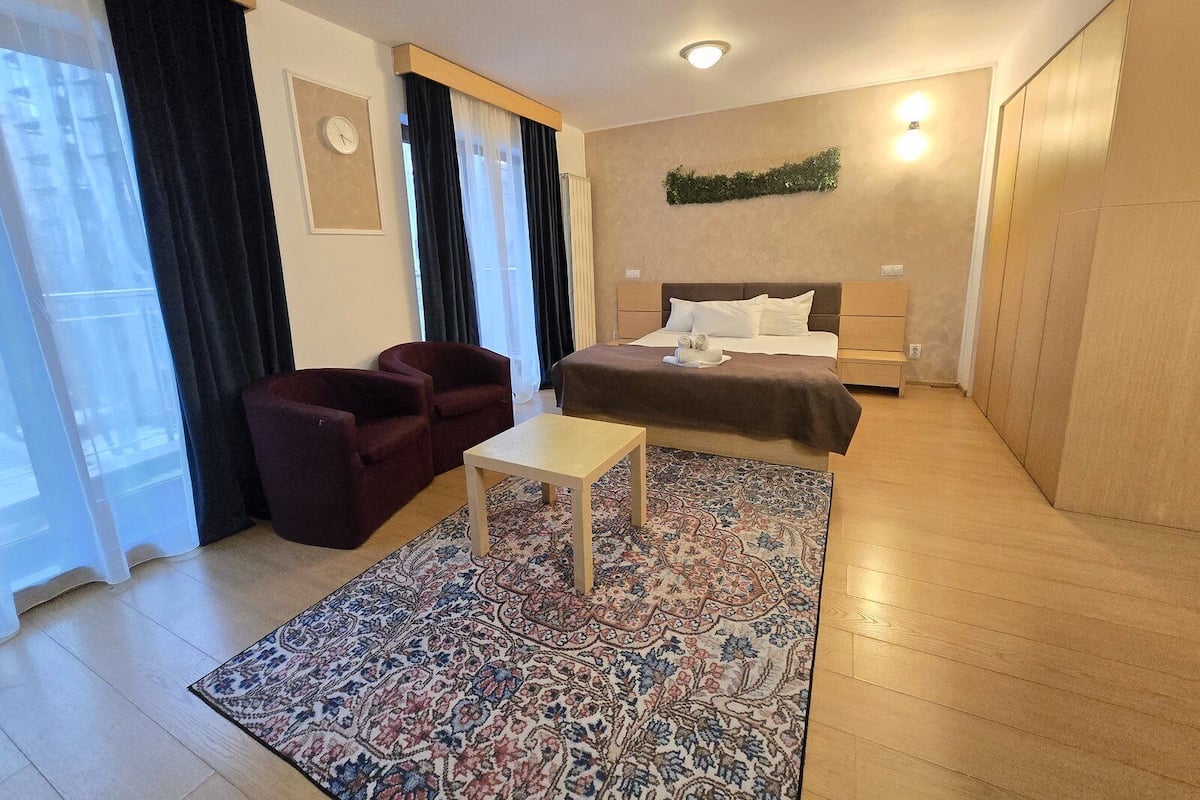 A hotel room with a bed, a small table, a chair, a patterned rug, a clock, and a window with curtains.
