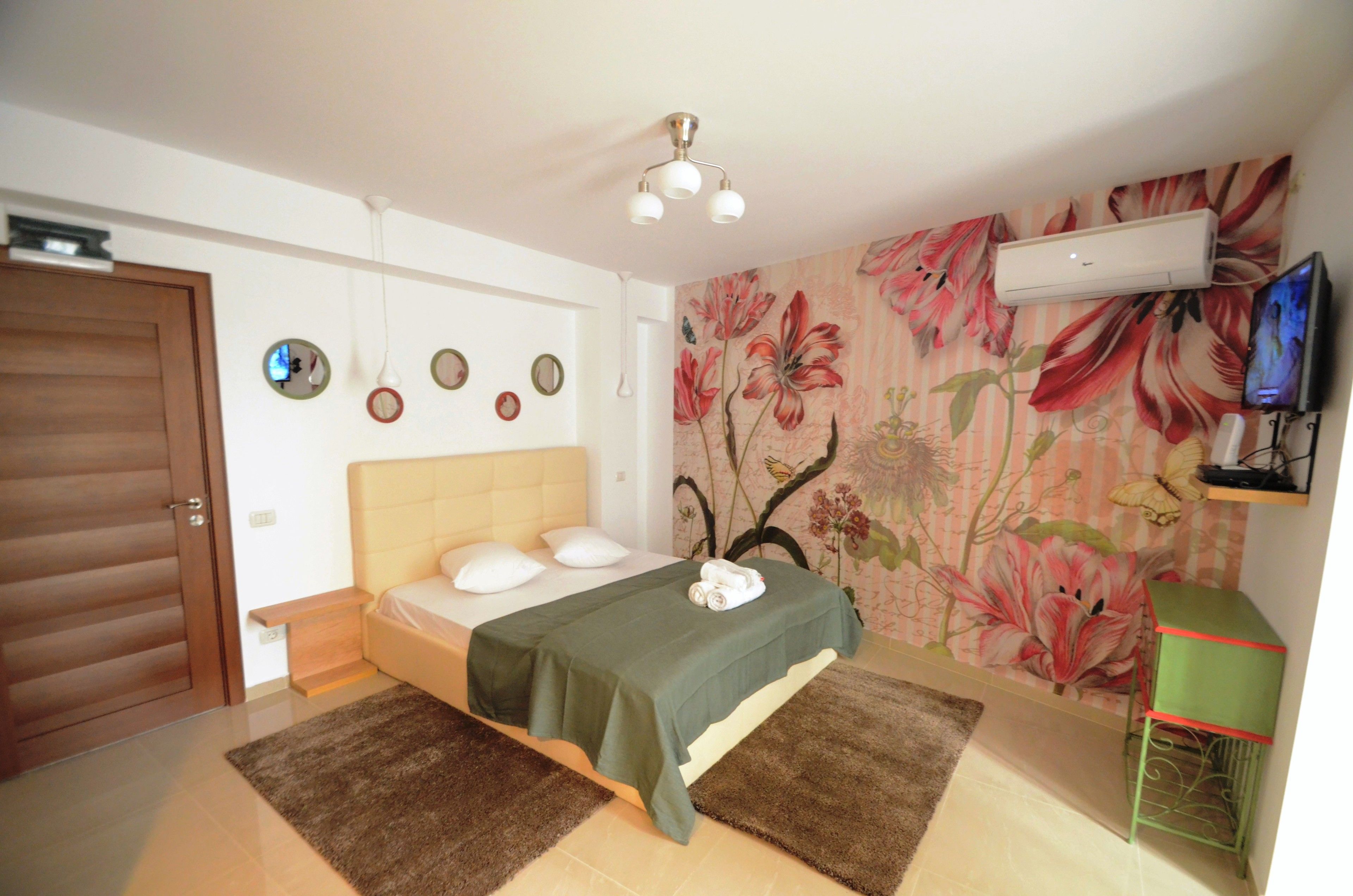 A bedroom with a bed, a green bedspread, a wall-mounted air conditioner, a television, and a floral wallpaper.