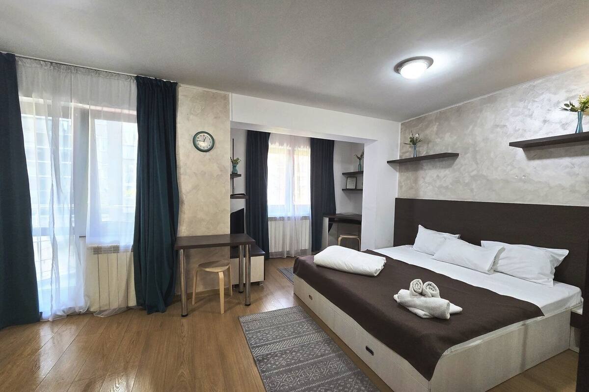 A hotel room with a bed, a desk, a chair, a clock, and a window with curtains.