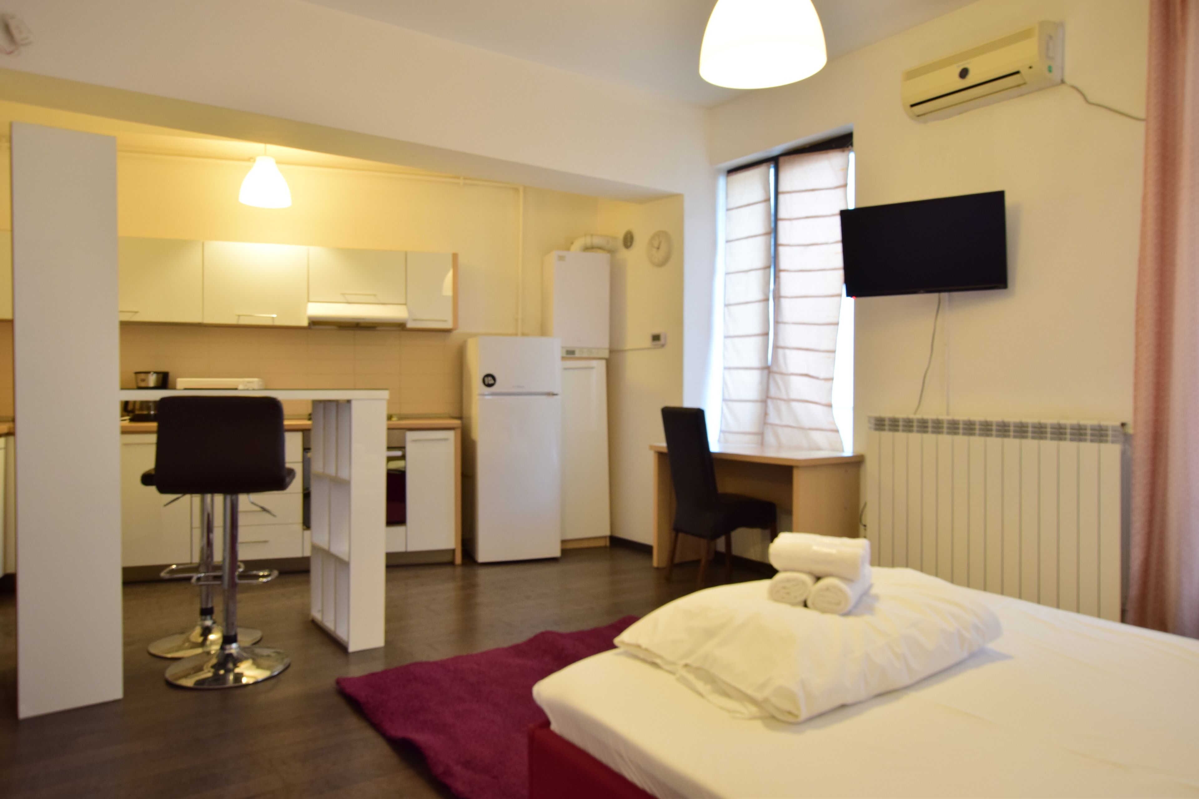 A compact hotel room with a bed, a desk, a chair, a small kitchenette, and a TV mounted on the wall.
