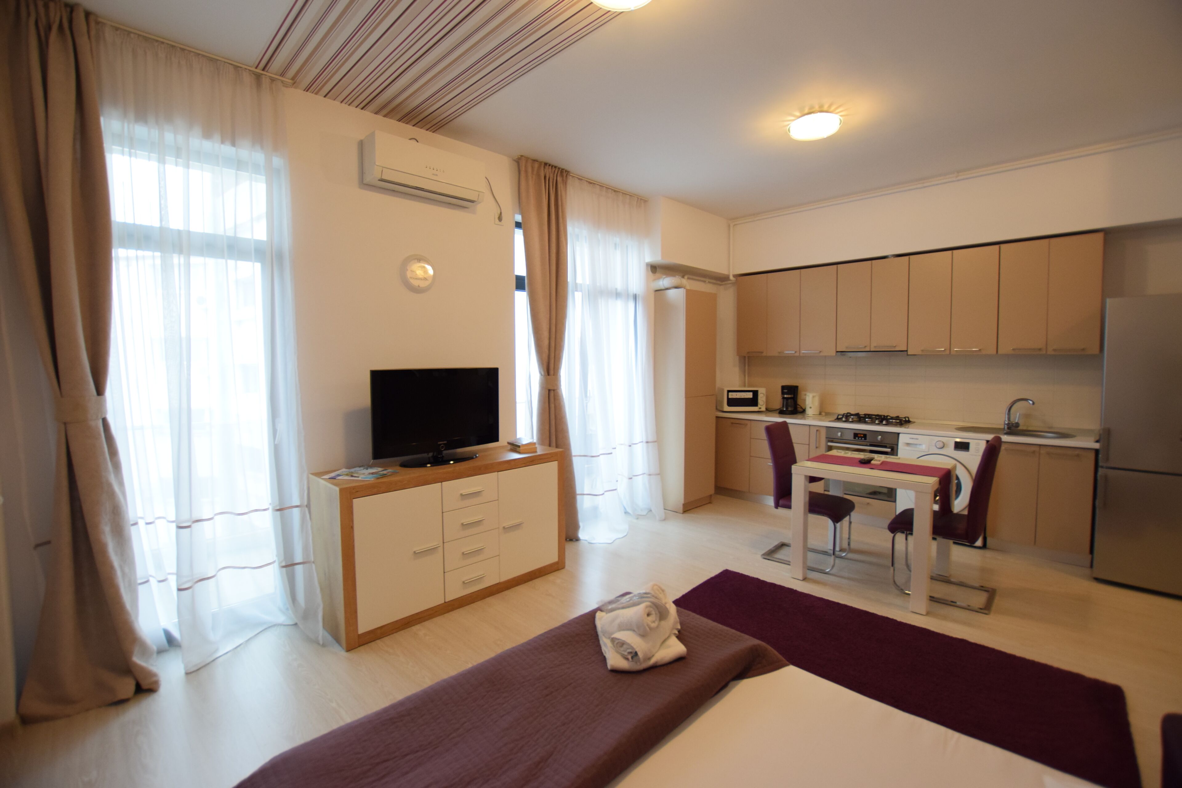 A modern hotel room with a bed, a TV, a desk with a chair, a kitchen area, and a bathroom.