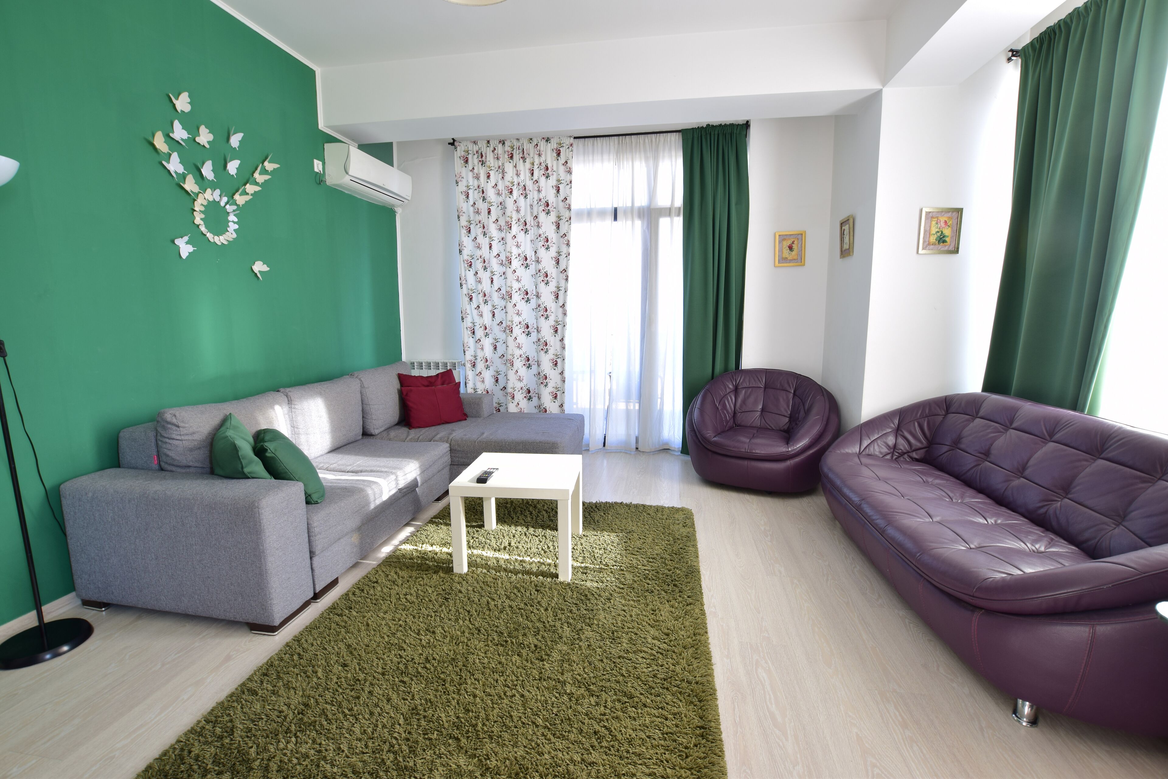 Een woonkamer met een grijze bank, een witte salontafel, een paarse fauteuil en een groen tapijt.