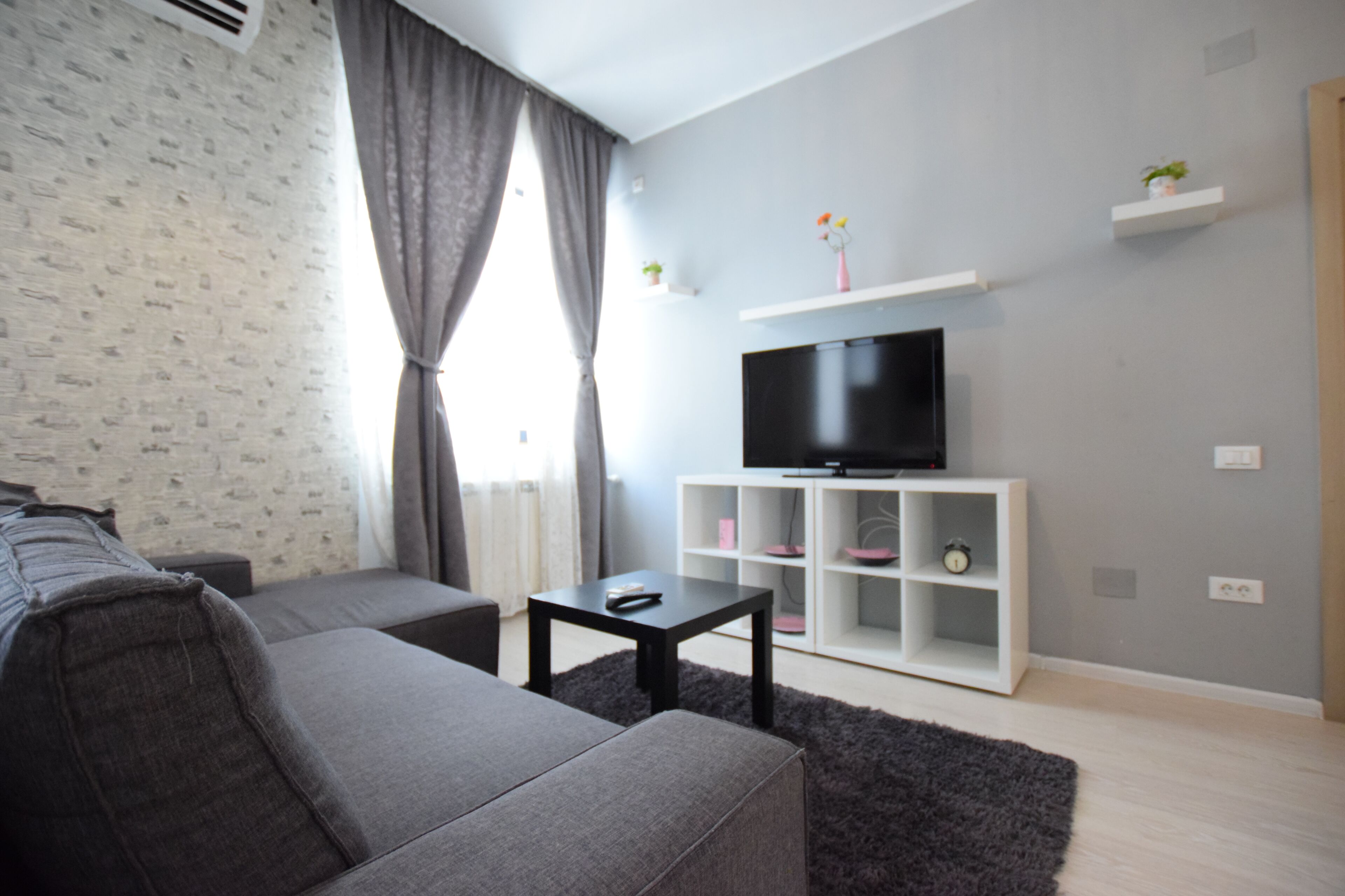 A modern living room with a grey sofa, a black coffee table, a flat-screen TV on a white stand, and a small white shelving unit with decorative items.