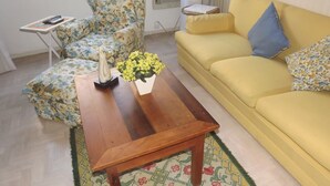 Een woonkamer met een gele bank, een fauteuil met bloemenprint, een houten salontafel met een vaas vol gele bloemen, en een klein bijzettafeltje met een boek.