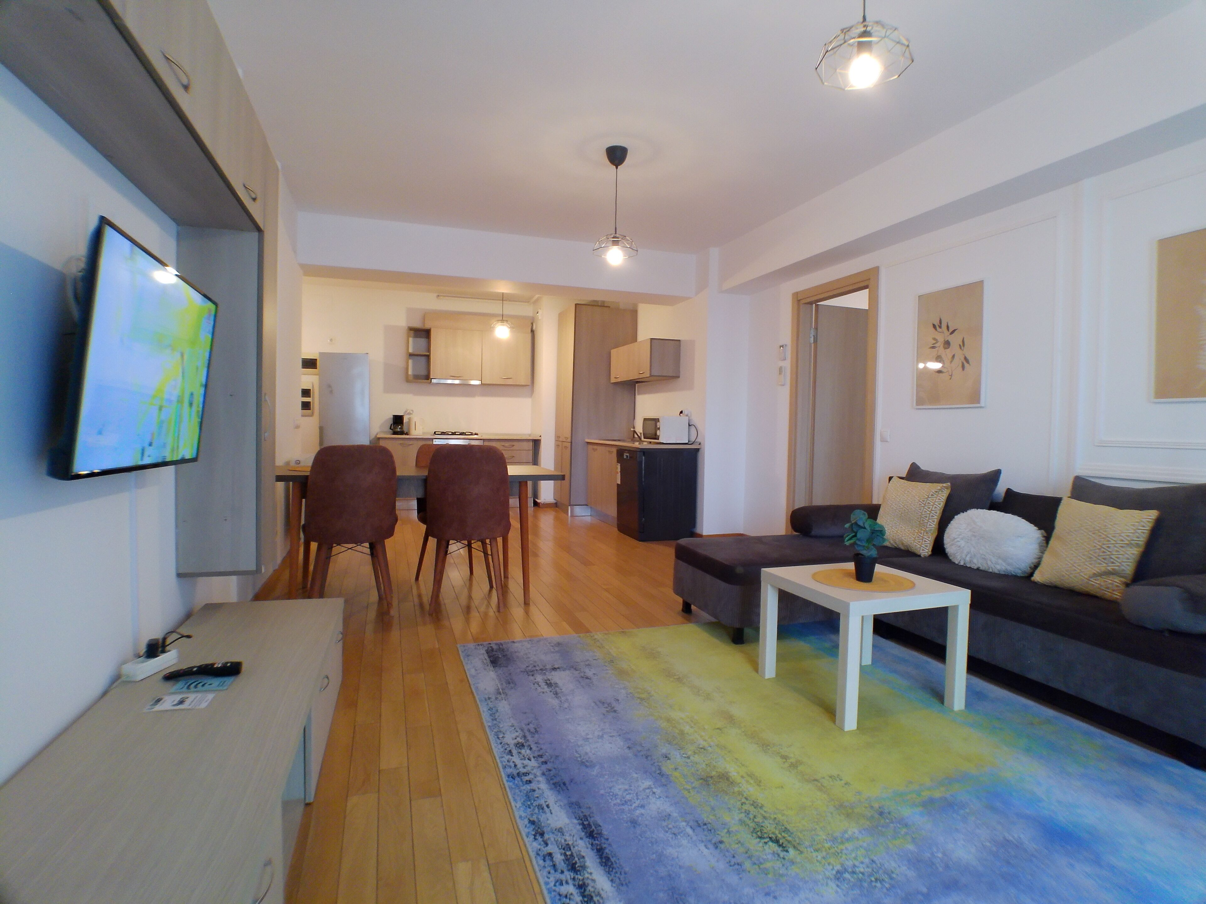 Un salon moderne avec un canapé, une table basse et un téléviseur. On aperçoit une cuisine à l’arrière-plan, avec des placards et un four à micro-ondes.