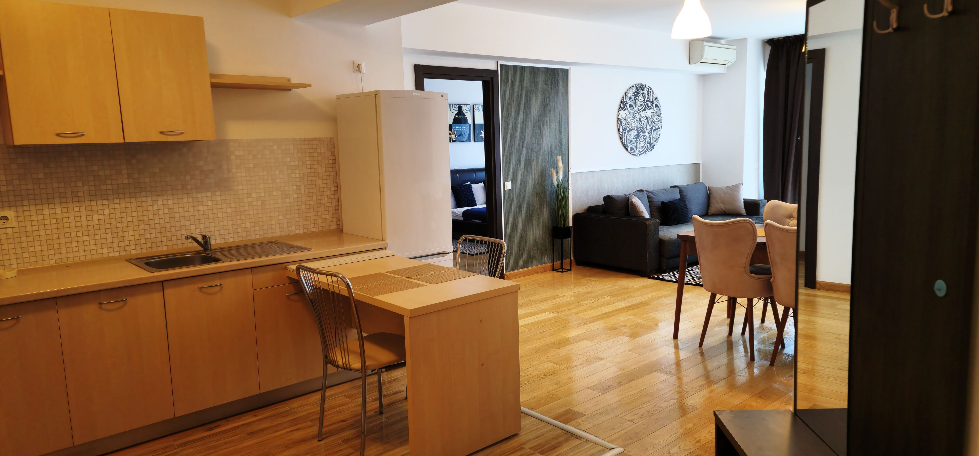 A compact kitchen with wooden cabinets and a tiled backsplash, a dining area with a table and chairs, and a living space with a sofa and a decorative wall piece.