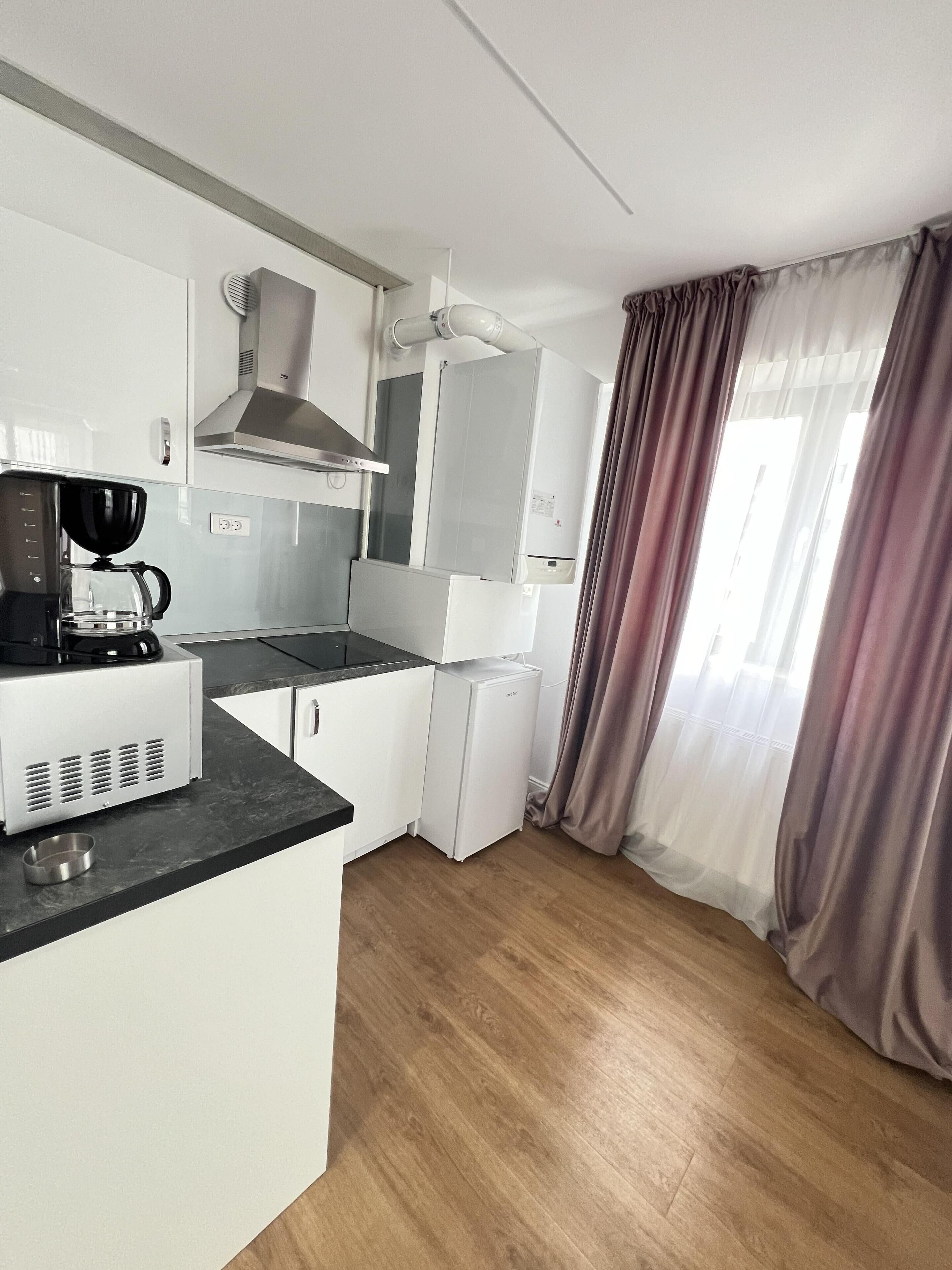 Cocina compacta con armarios blancos, cafetera, refrigerador pequeño y ventana con cortinas.
