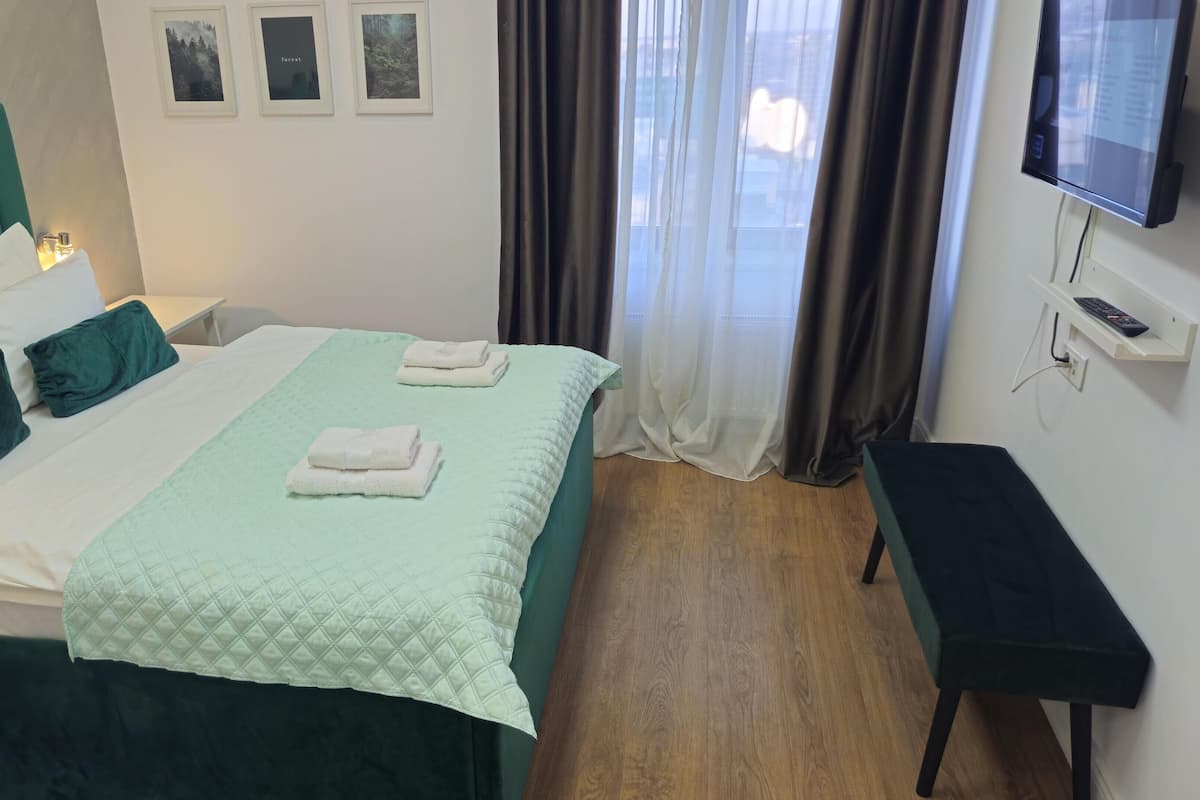 Una cama bien hecha con colcha verde acolchada, dos almohadas y toallas dobladas. Hay una mesita de noche con una lámpara, un televisor montado en la pared y una ventana con cortinas.