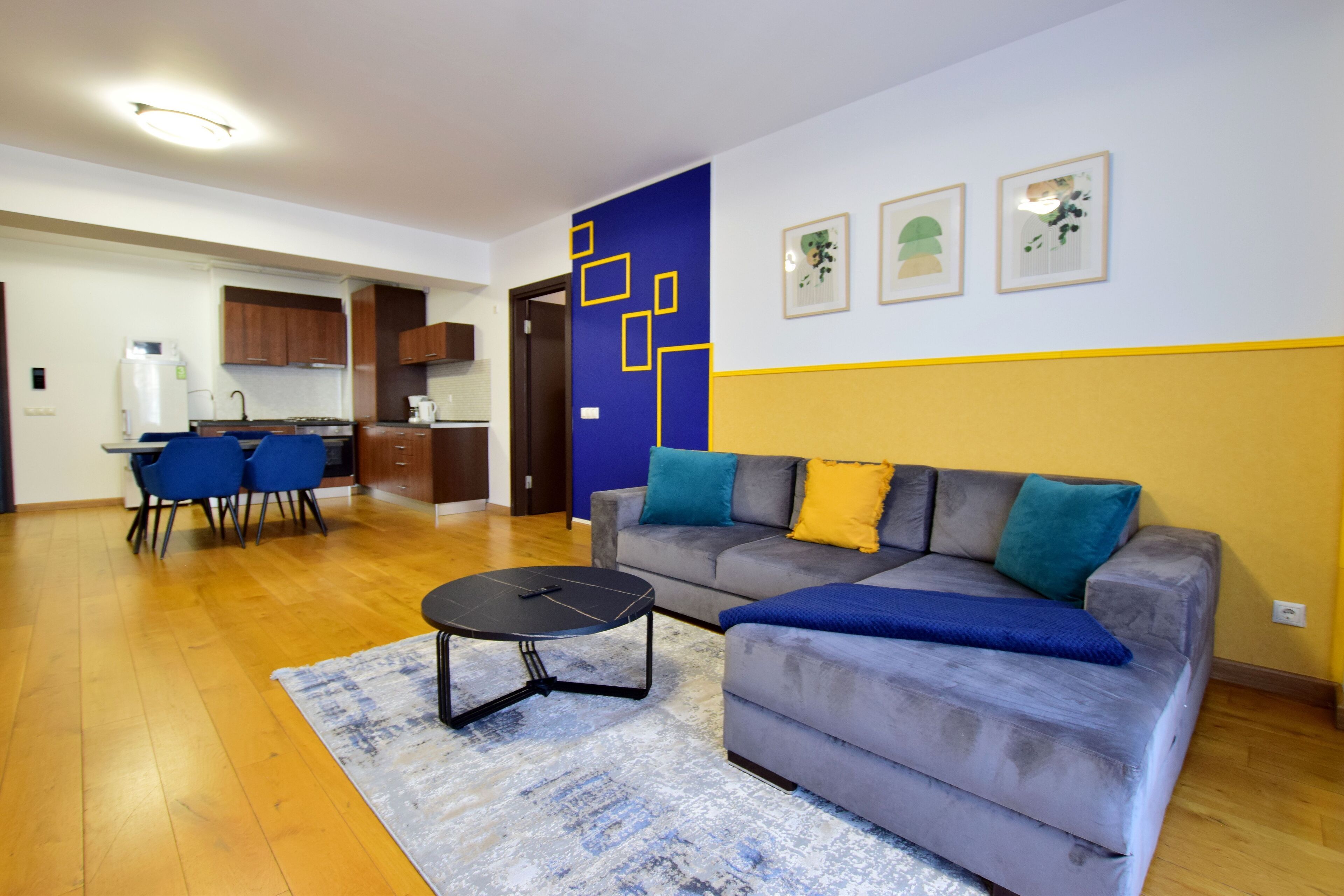 A modern living room with a grey sofa, blue and yellow cushions, a black coffee table, and a blue rug. There is a kitchen area with wooden cabinets and a dining table with blue chairs.
