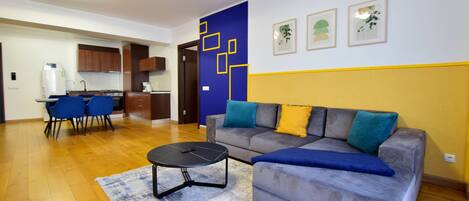 A modern living room with a grey sofa, blue and yellow cushions, a black coffee table, and a blue rug. There is a kitchen area with wooden cabinets and a dining table with blue chairs.