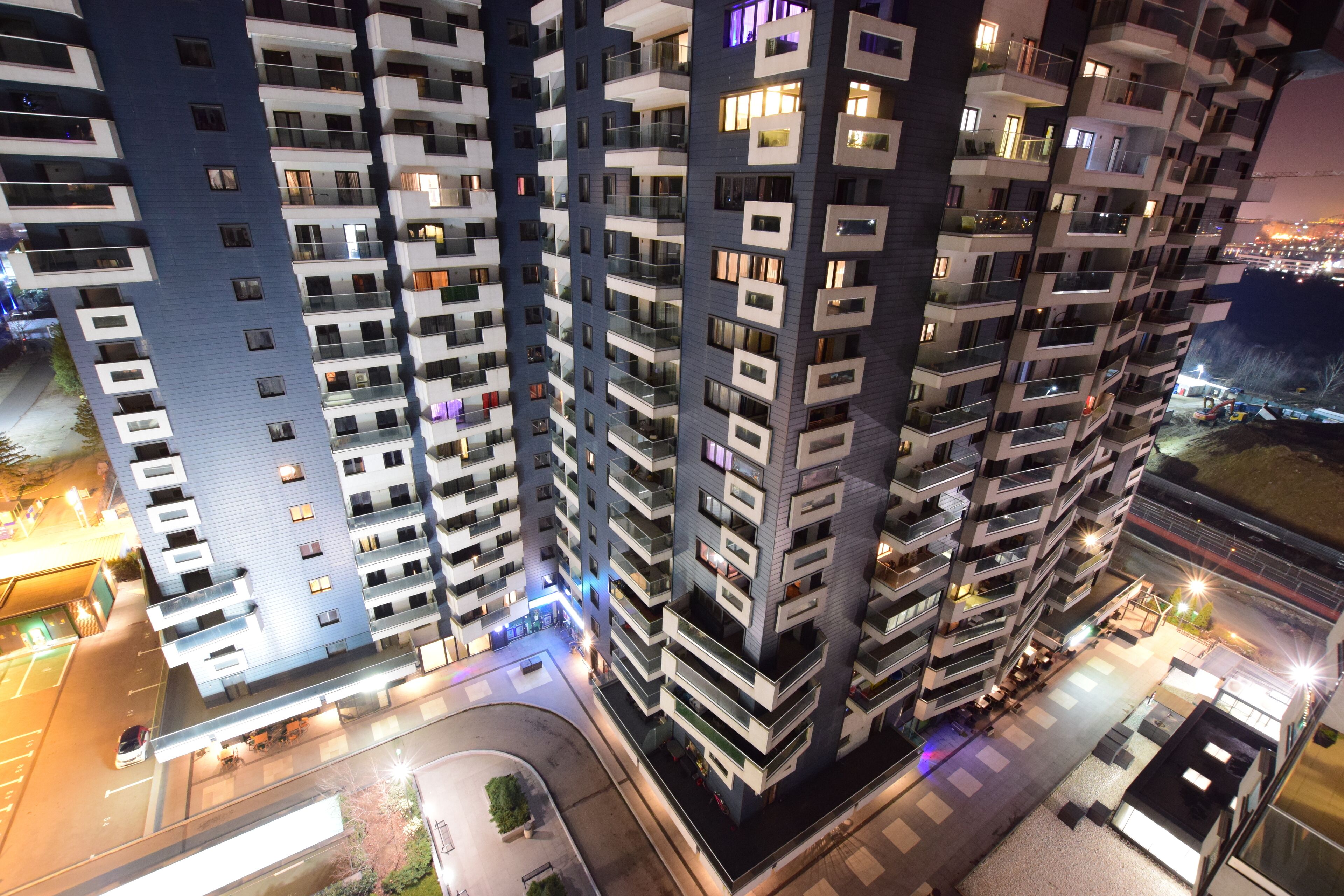 A high-rise residential building with multiple balconies, a parking area, and a well-lit street at night.