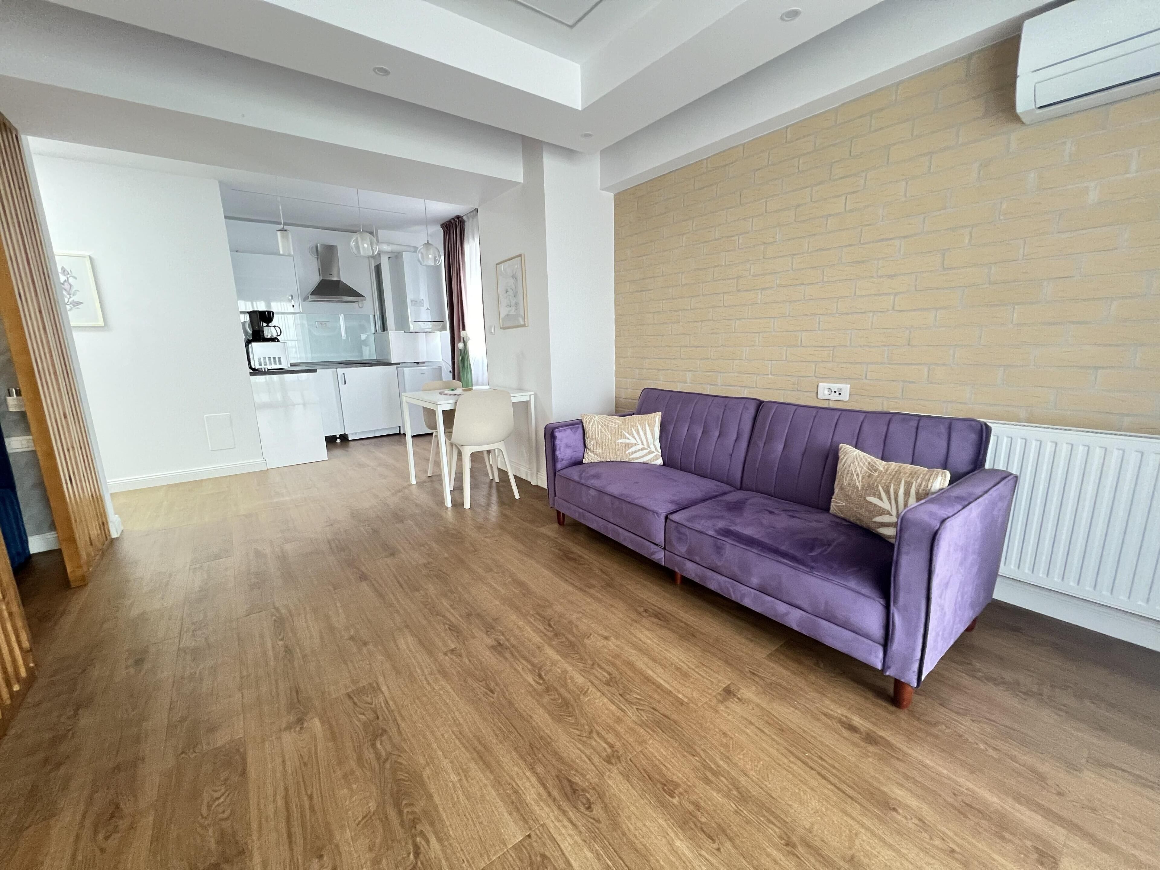 A modern living room with a purple sofa, a dining table with chairs, and a kitchen in the background.