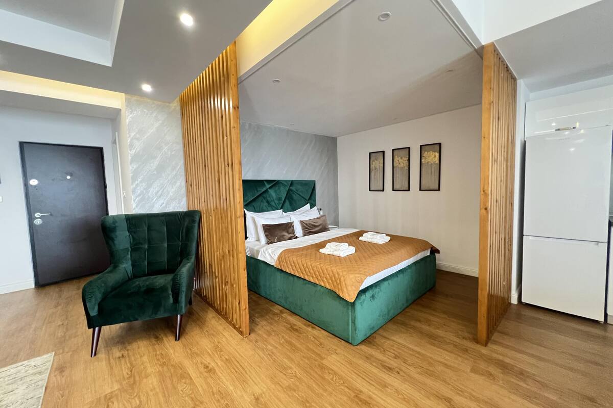 Una habitación de hotel moderna con una cama tapizada en verde, una pared de acento de madera y un refrigerador blanco.