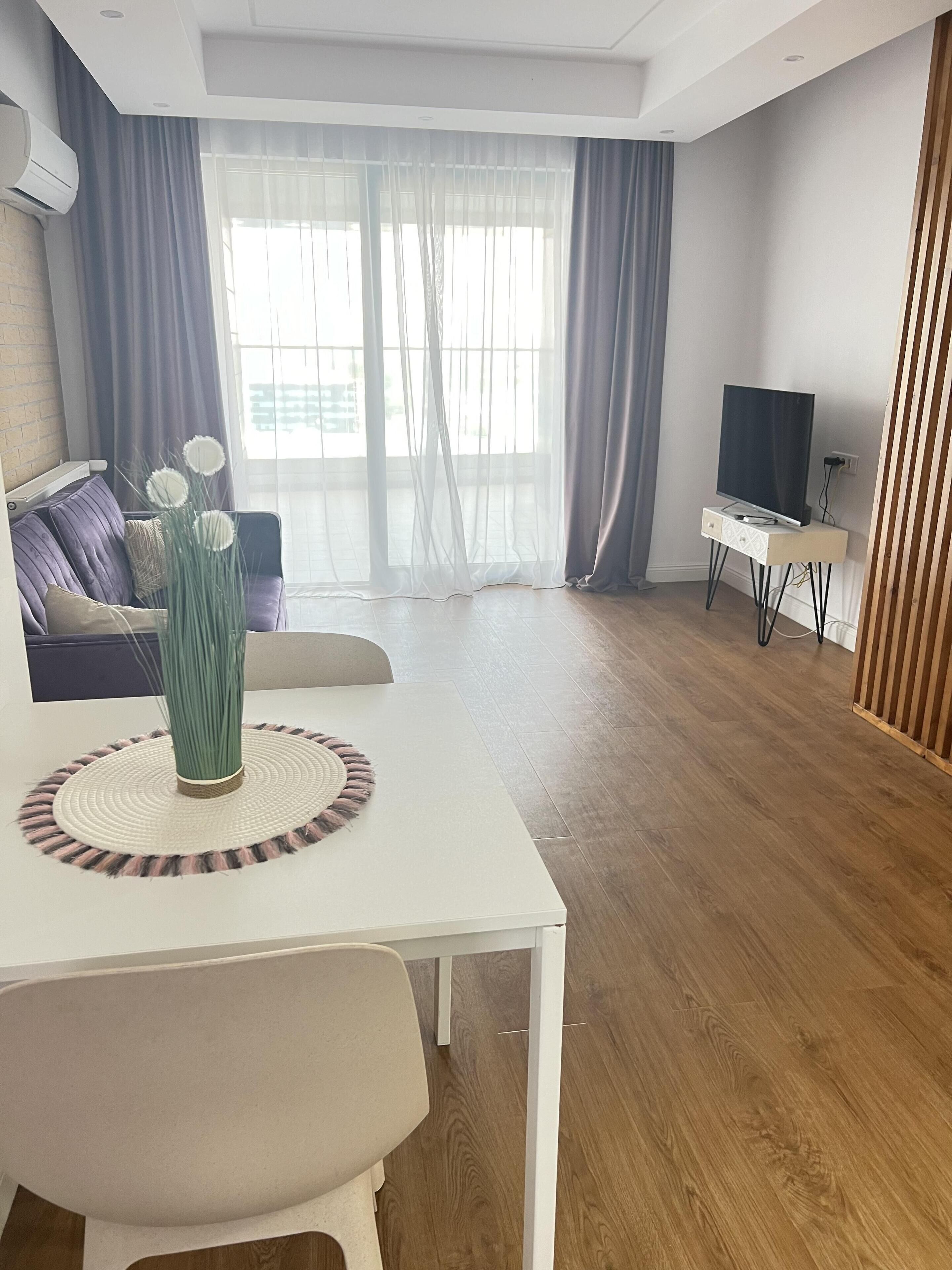 Ein modernes Wohnzimmer mit einem weißen Tisch, einer Couch, einem Fernseher auf einem Standfuß und einem großen Fenster mit Vorhängen.