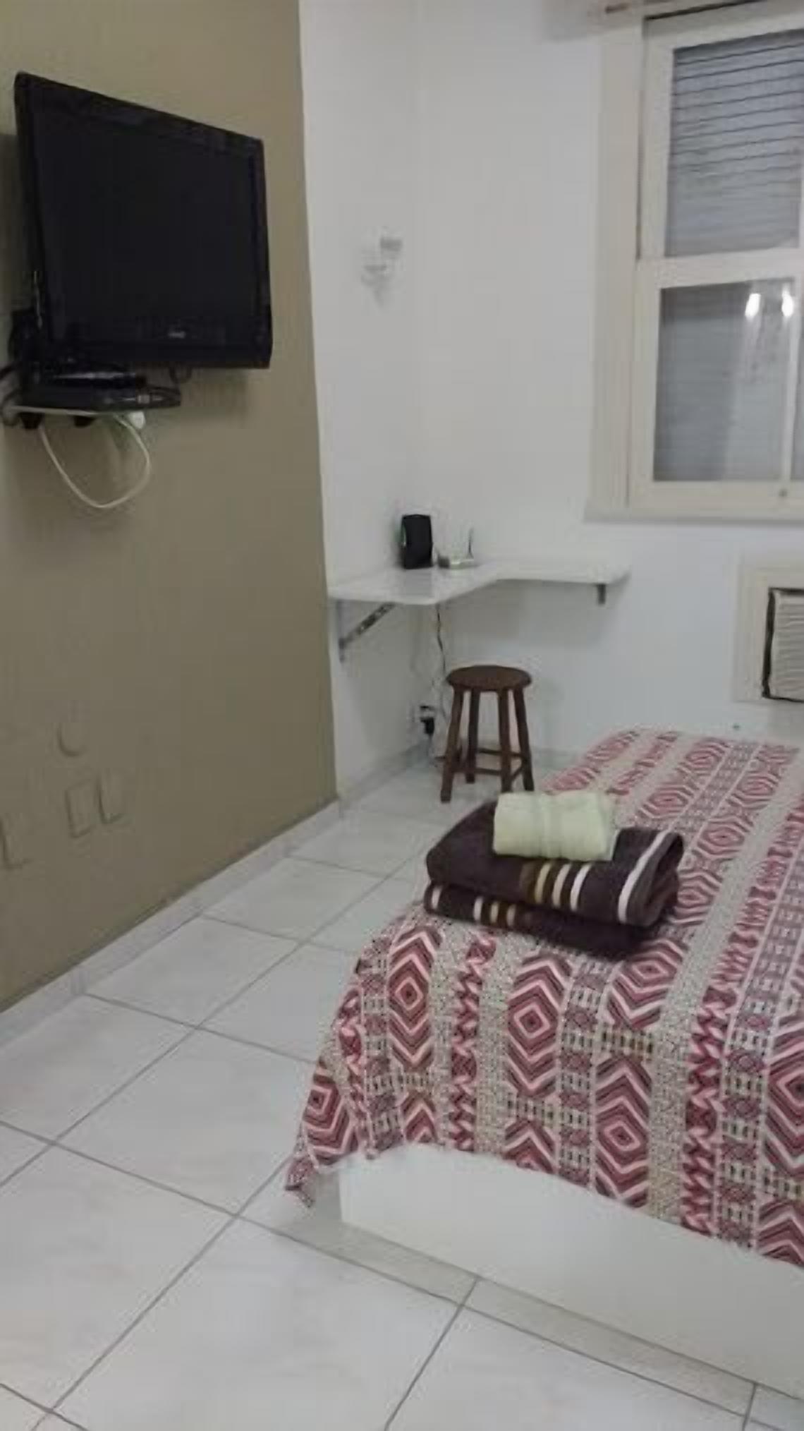 A bedroom with a bed, a television mounted on the wall, a small stool, and a window with blinds.