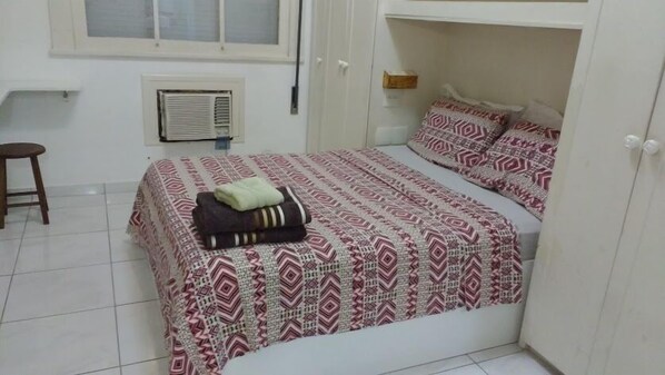 A bedroom with a bed, a window, an air conditioning unit, a stool, and a wardrobe.