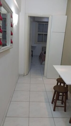 A narrow hallway with a white tiled floor leading to a room with a window. There is a white refrigerator on the right and a wooden stool in the foreground.
