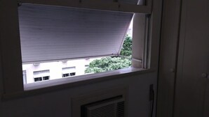 Una ventana con una persiana blanca, medio bajada, muestra un edificio y follaje al exterior.