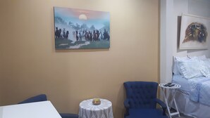 A room with a painting of horses, a blue armchair, a small round table with a candle, and a bed with white bedding. - Kitnet Hilário de Gouveia (Rio de Janeiro)