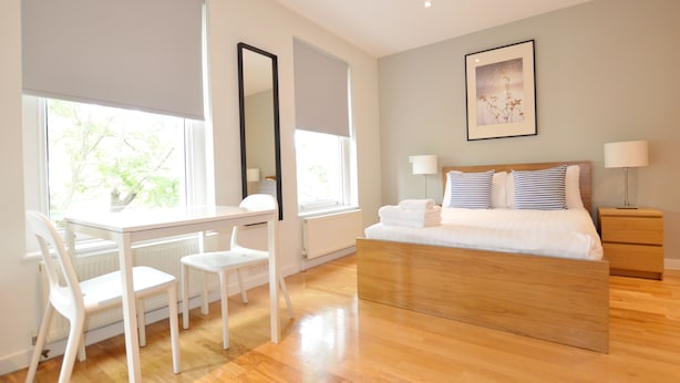 Um quarto moderno com uma cama de madeira, uma escrivaninha com cadeiras, janelas amplas e um quadro emoldurado na parede.