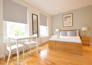 A modern bedroom with a wooden bed, a desk with chairs, large windows, and a framed picture on the wall. - Russell Square Serviced Apartments by Concept Apartments (London)