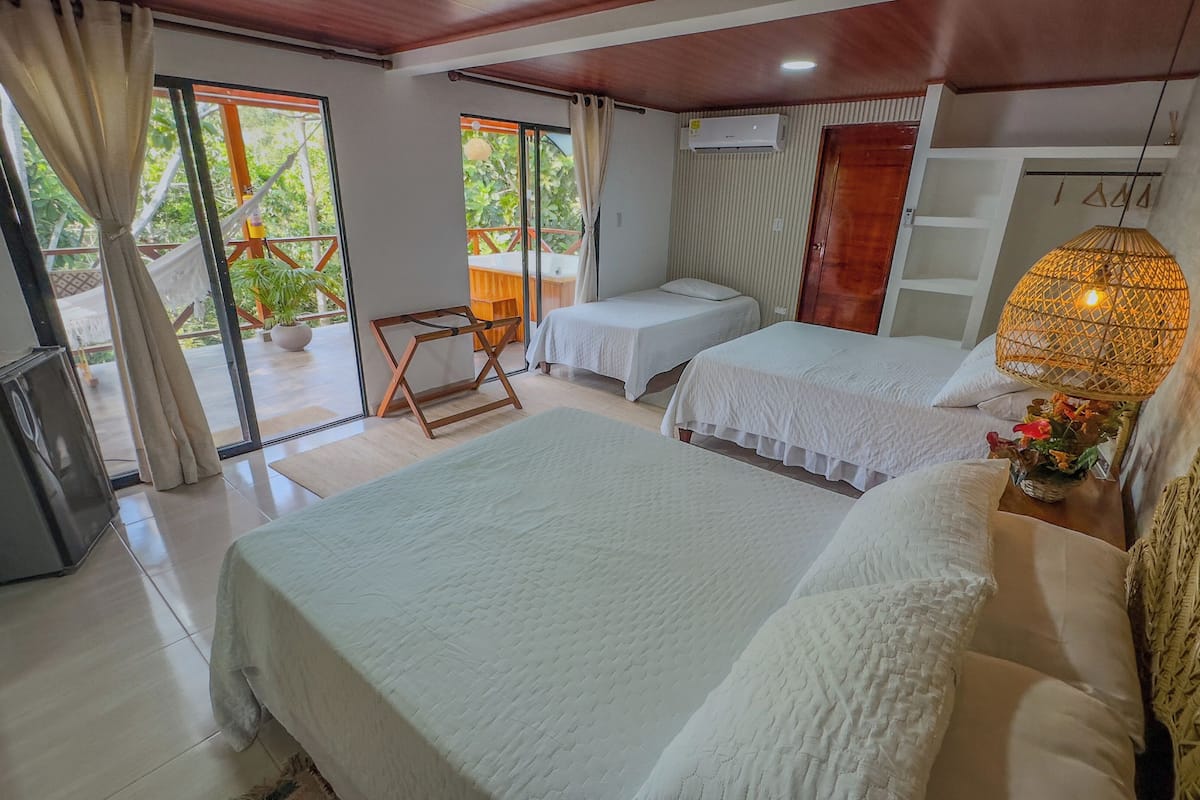 A bedroom with two beds, a small desk, a chair, and a balcony with a view of greenery.