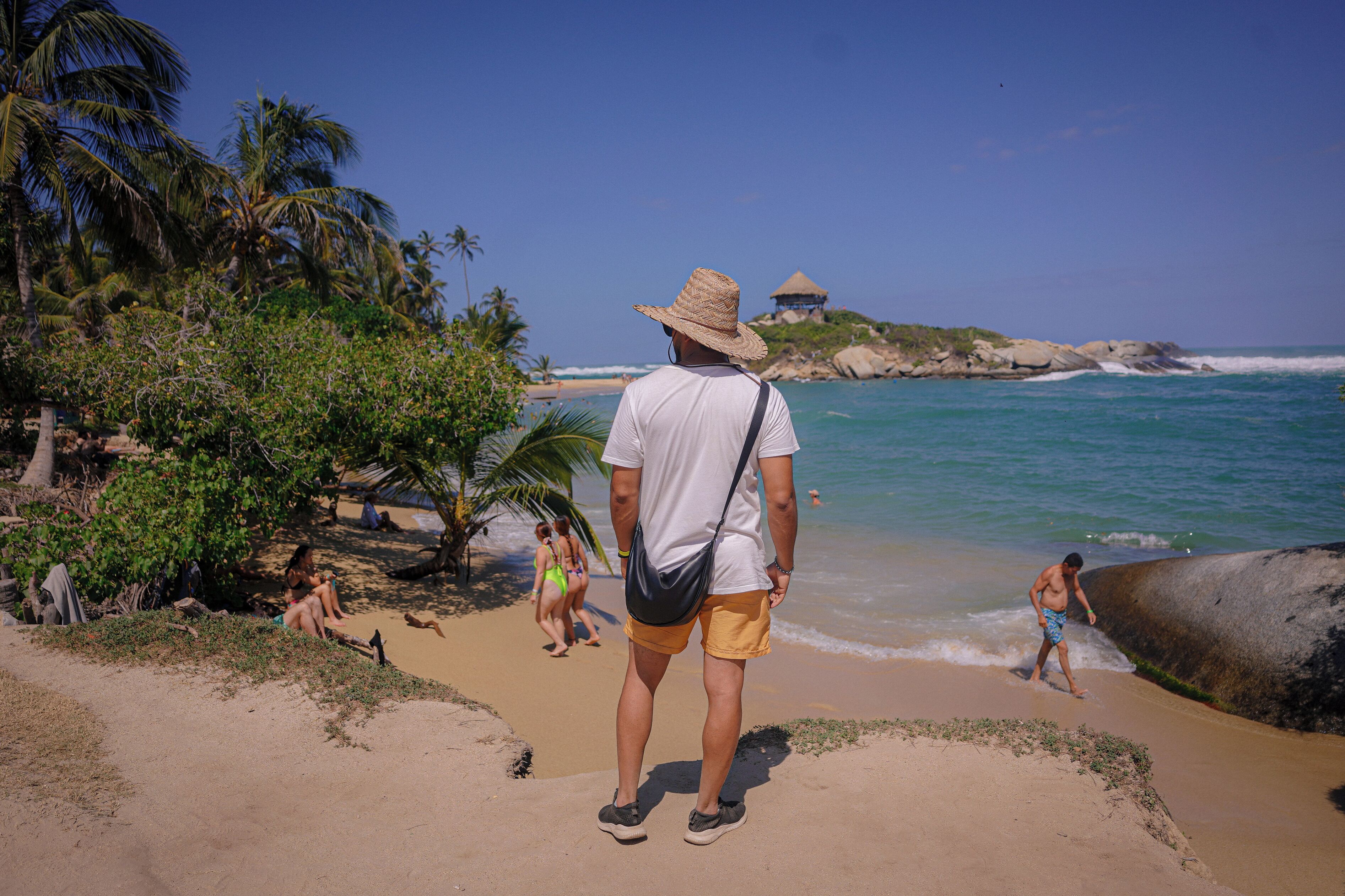 Escena de playa con un hombre de sombrero de paja y pantalones cortos, palmeras y una cabaña al fondo.