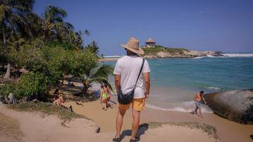 Escena de playa con un hombre de sombrero de paja y pantalones cortos, palmeras y una cabaña al fondo.