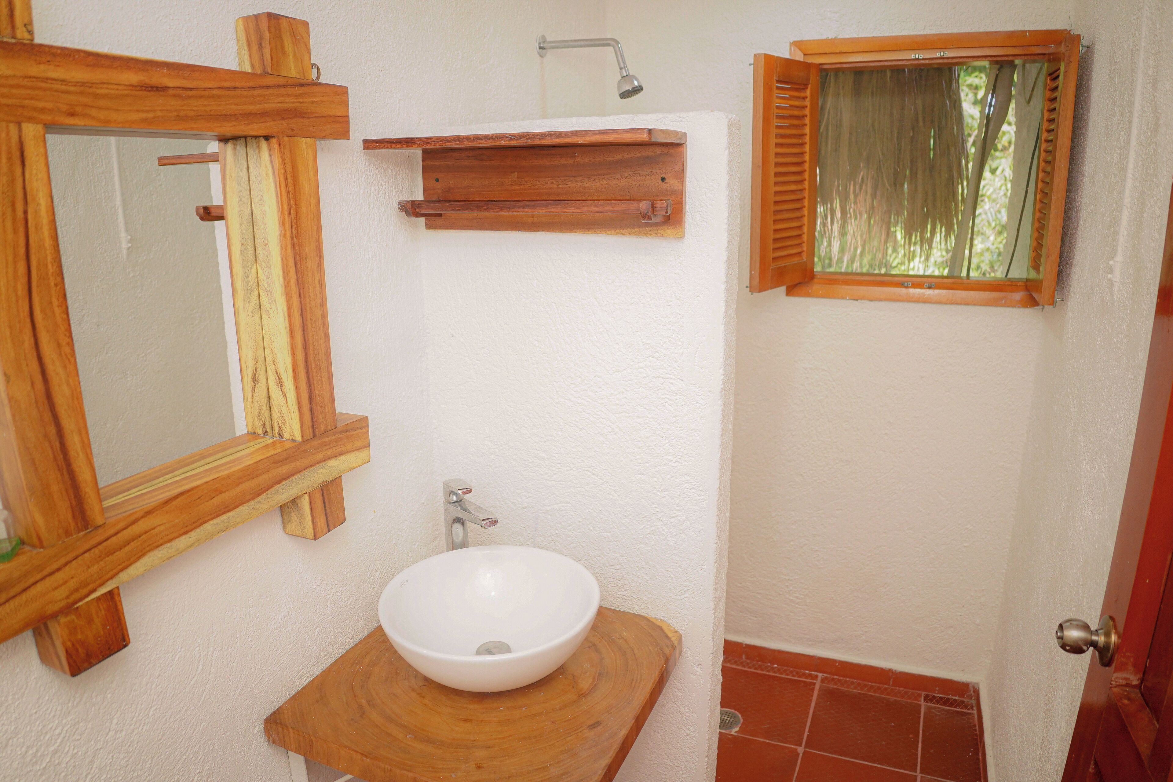Un baño con tocador de madera, lavamanos blanco, espejo, ducha y ventana con contraventanas.