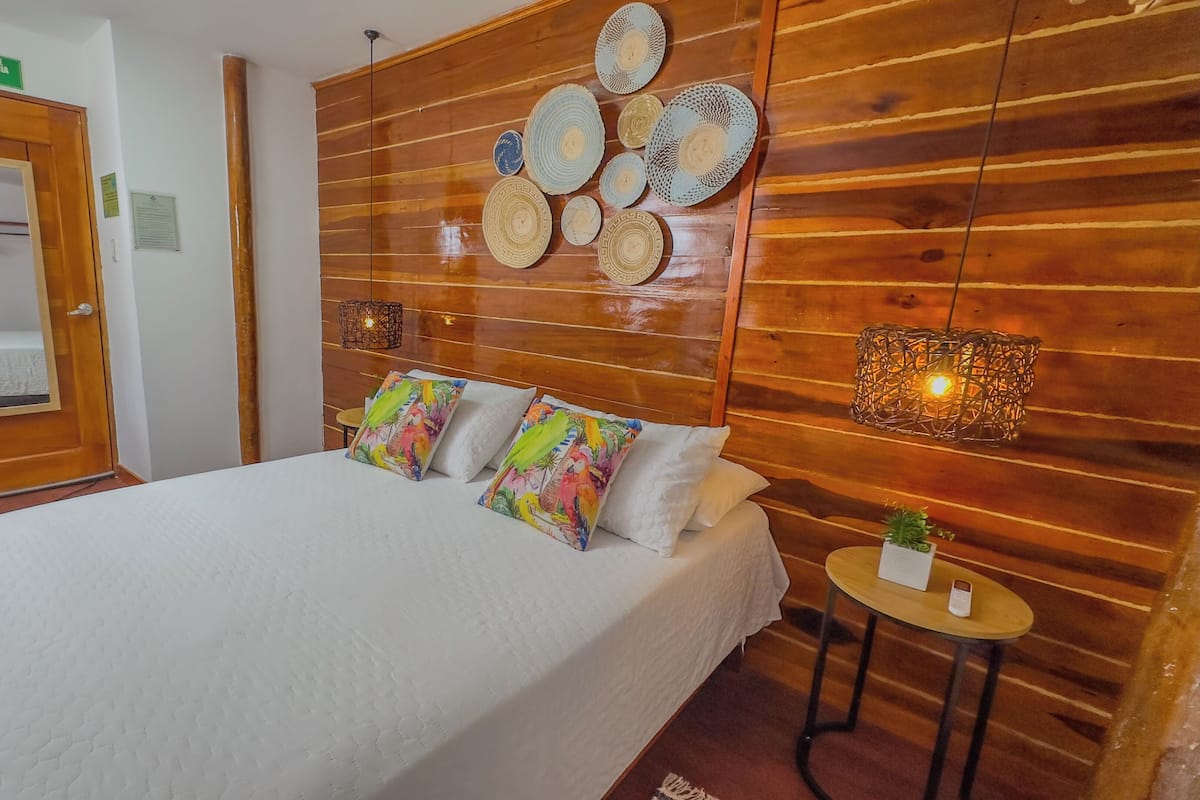 A hotel room with a wooden headboard, a bed with white bedding, colorful pillows, a small table with a plant, and a wall-mounted lamp.