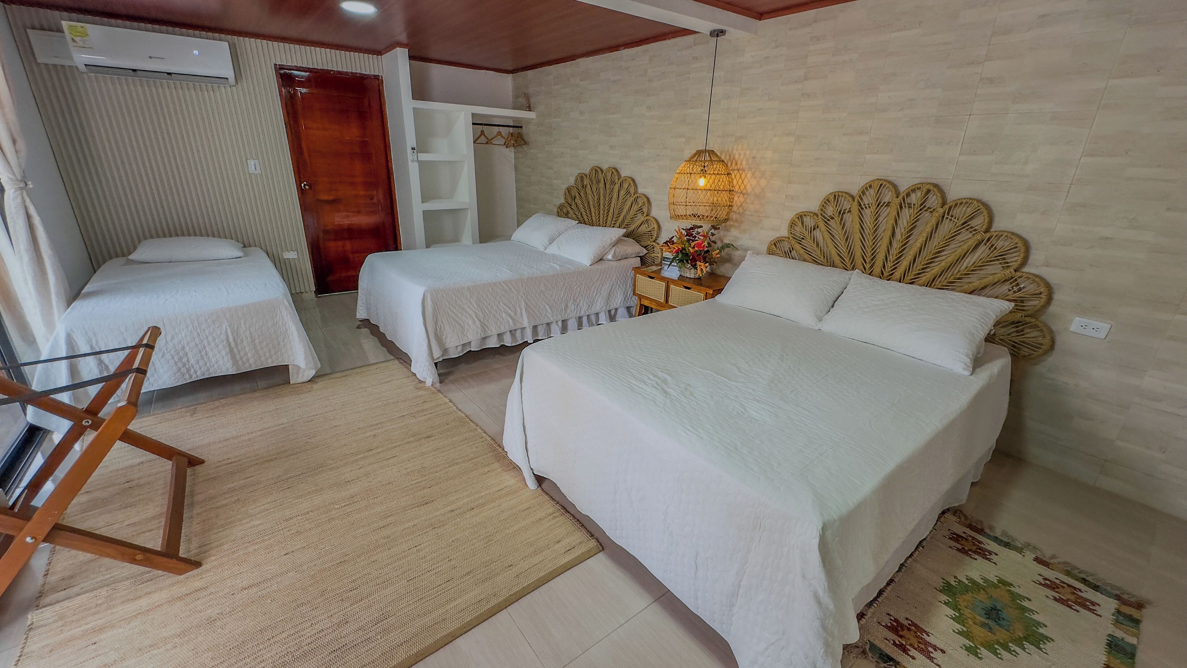 A room with a large bed, two smaller beds, a wooden chair, a woven headboard, a hanging light fixture, and a small table with a vase of flowers.