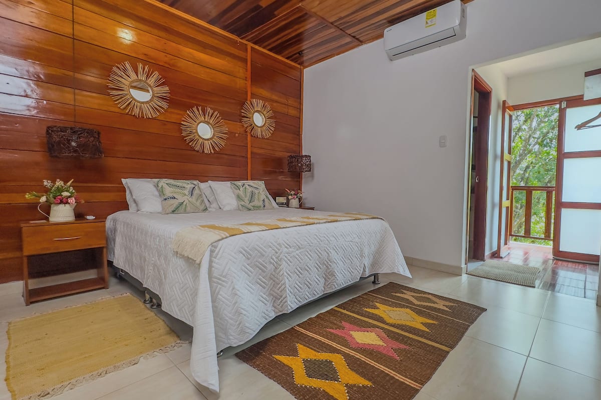 A bedroom with a wooden ceiling, a bed with a white quilt, a yellow rug, and a view of greenery through a door.