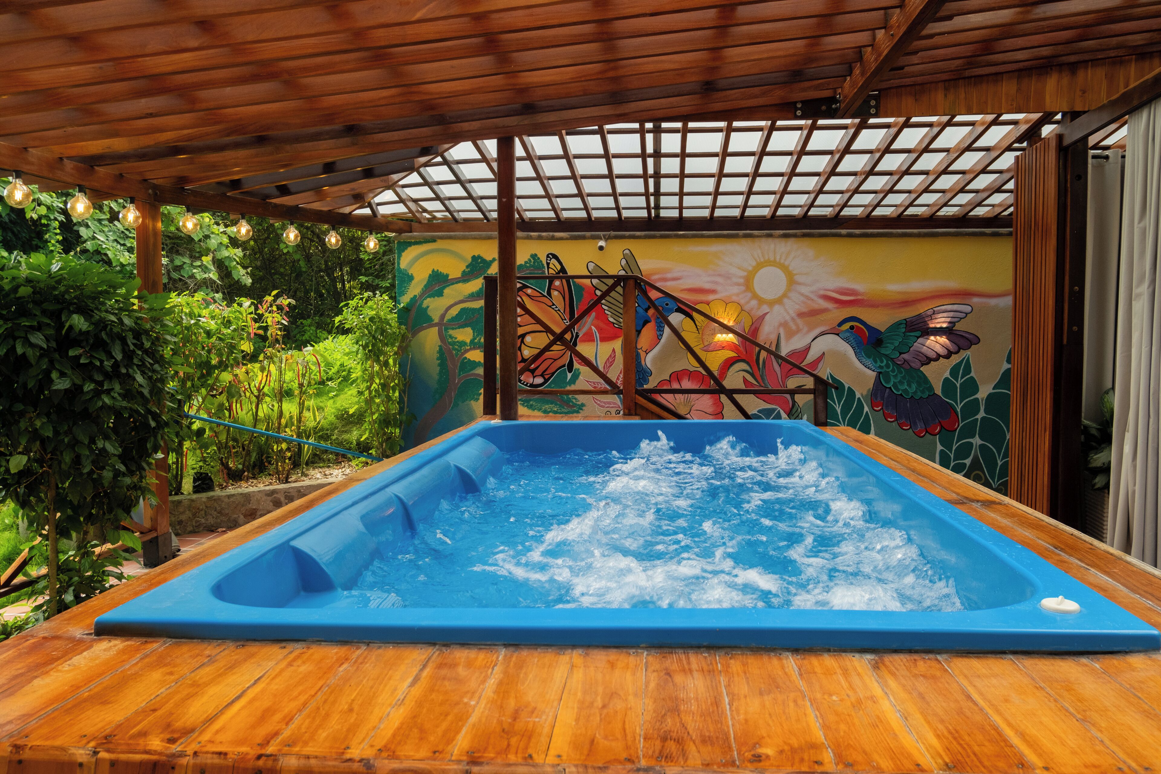 A wooden deck with a hot tub, surrounded by lush greenery and a colorful mural.