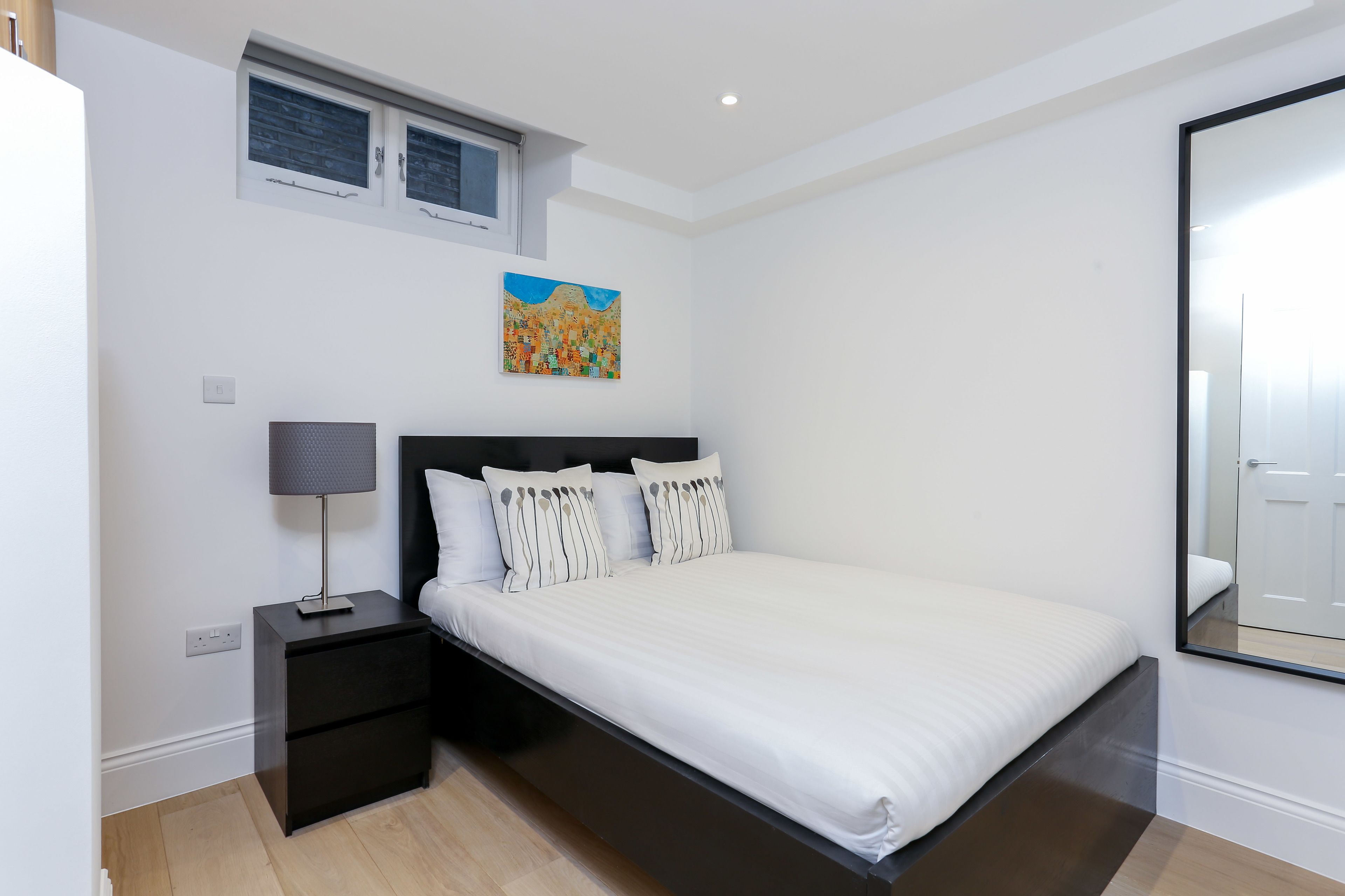Une chambre à coucher moderne avec un grand lit, une table de chevet, une lampe, une fenêtre et un tableau au mur.