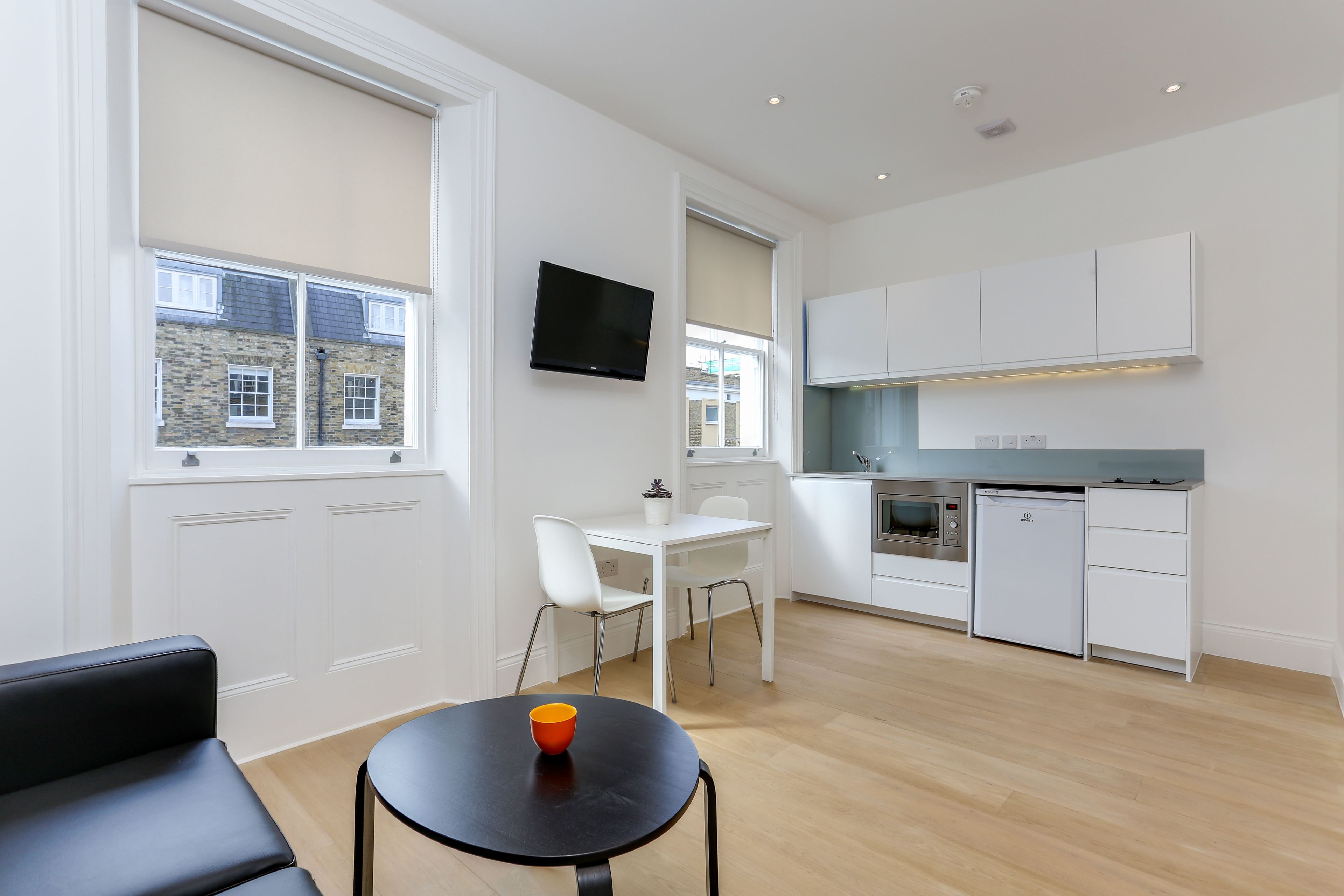 A modern kitchen with white cabinets, a built-in oven, and a microwave. There is a dining area with a white table and chairs, and a black sofa.