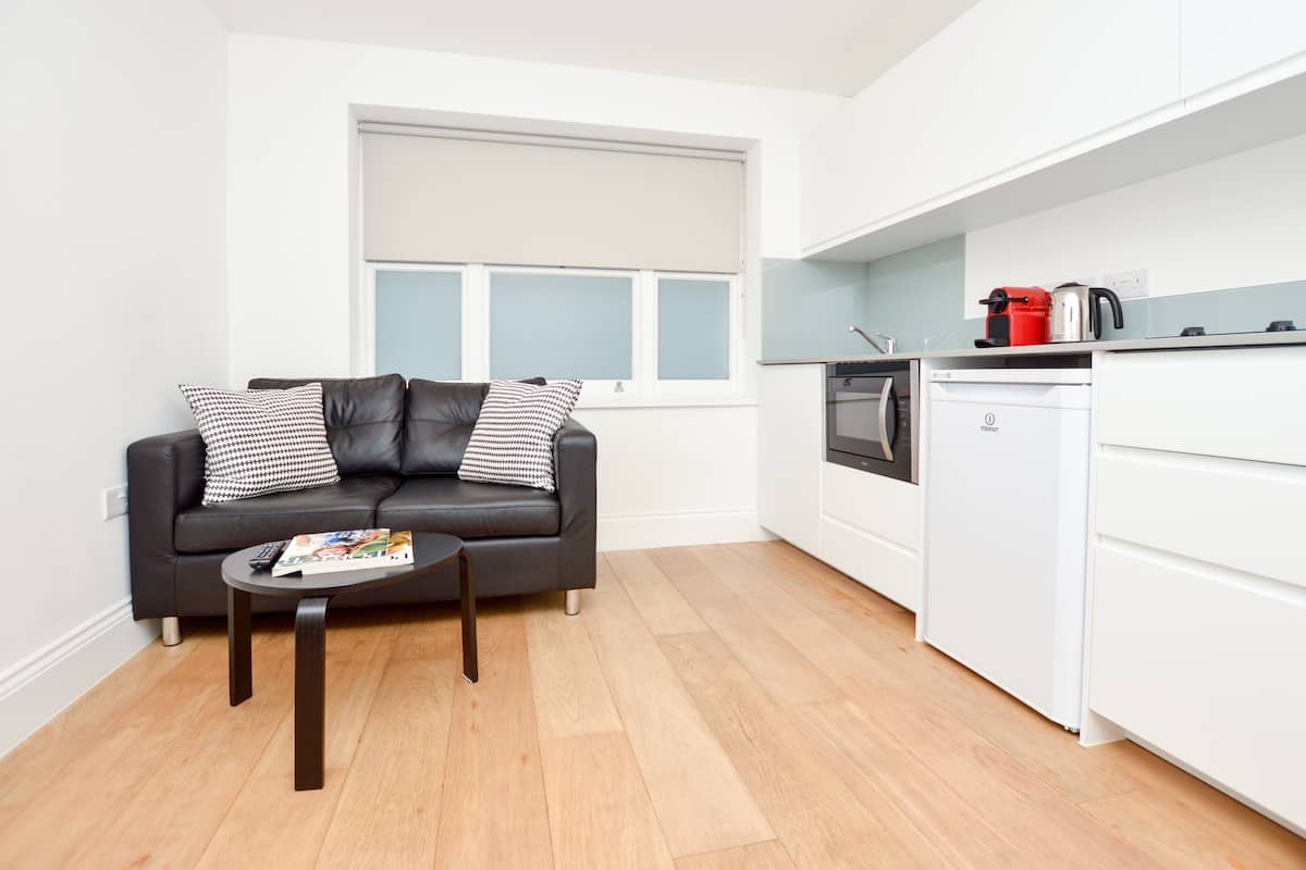 A compact living space with a black leather sofa, a small coffee table, a kitchen area with white cabinets, and a window with blinds.