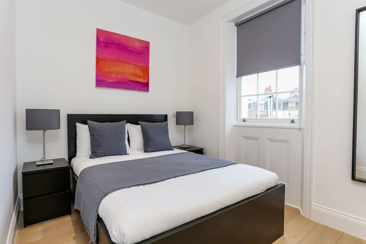 A neatly arranged bedroom with a large bed, two bedside tables, a window with a view, and a colorful abstract painting on the wall.