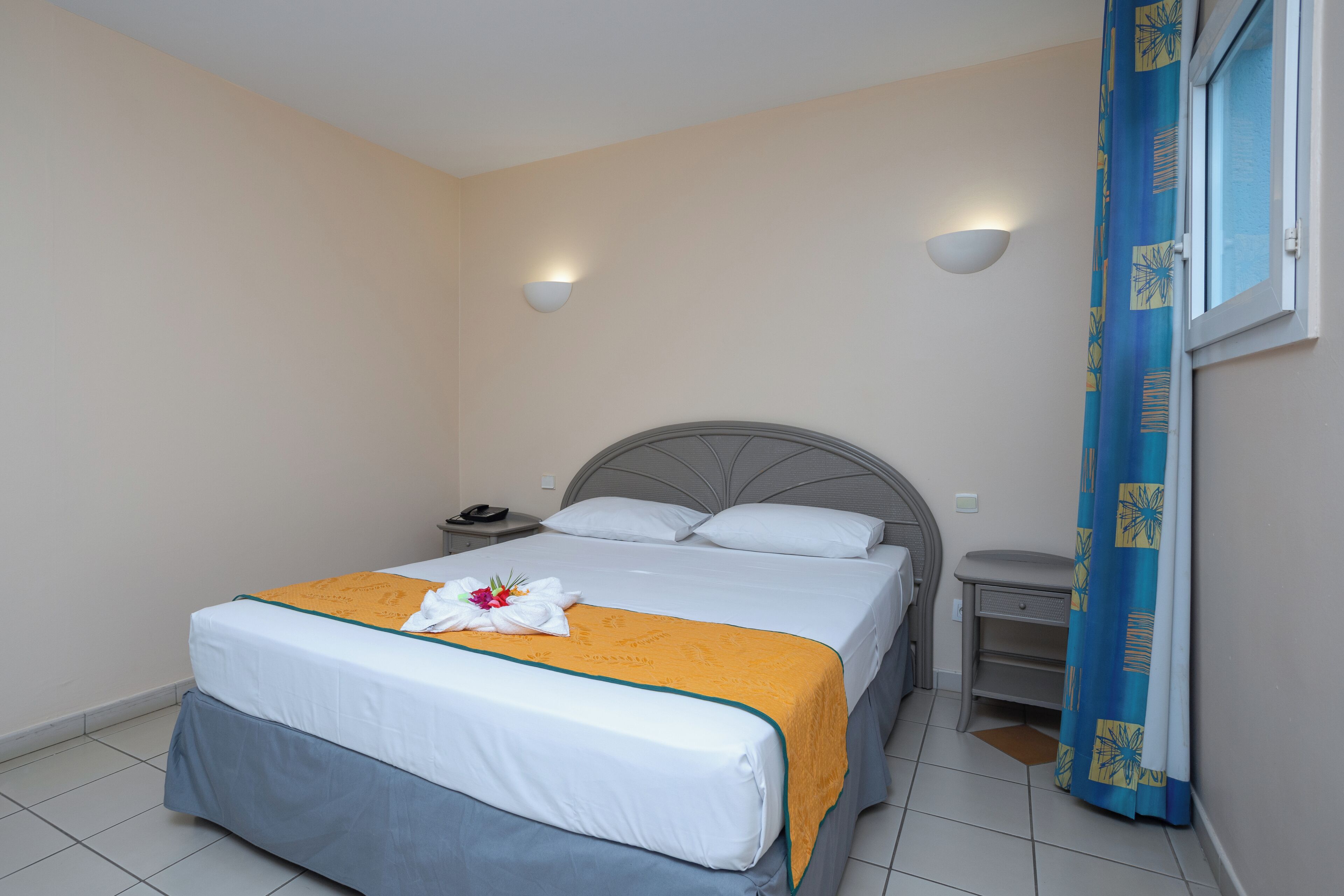 Een netjes opgemaakt bed met een wit en geel sprei, een nachtkastje met een telefoon, en een wandlamp.