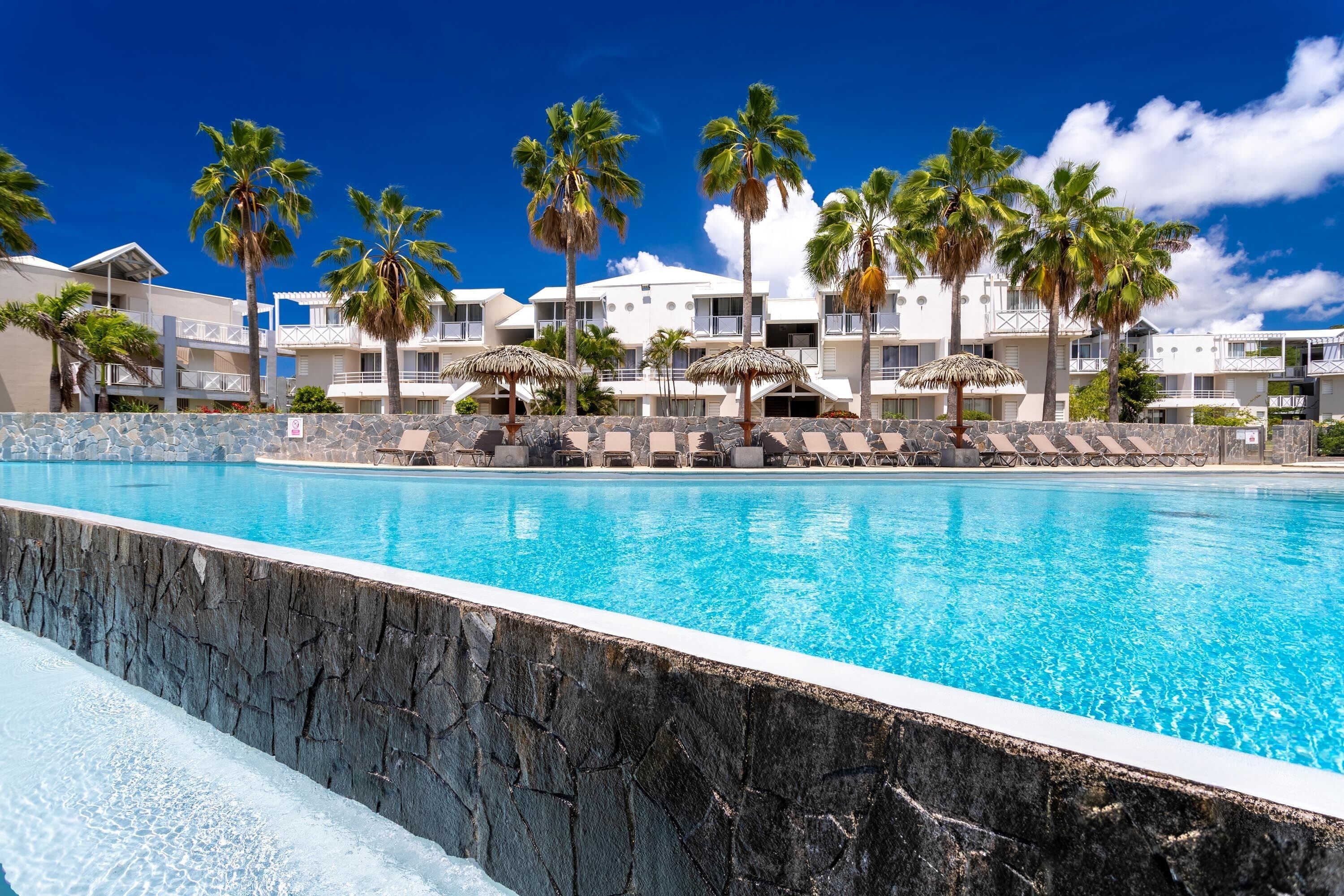 A swimming pool with a stone border, palm trees, and a building with balconies in the background.