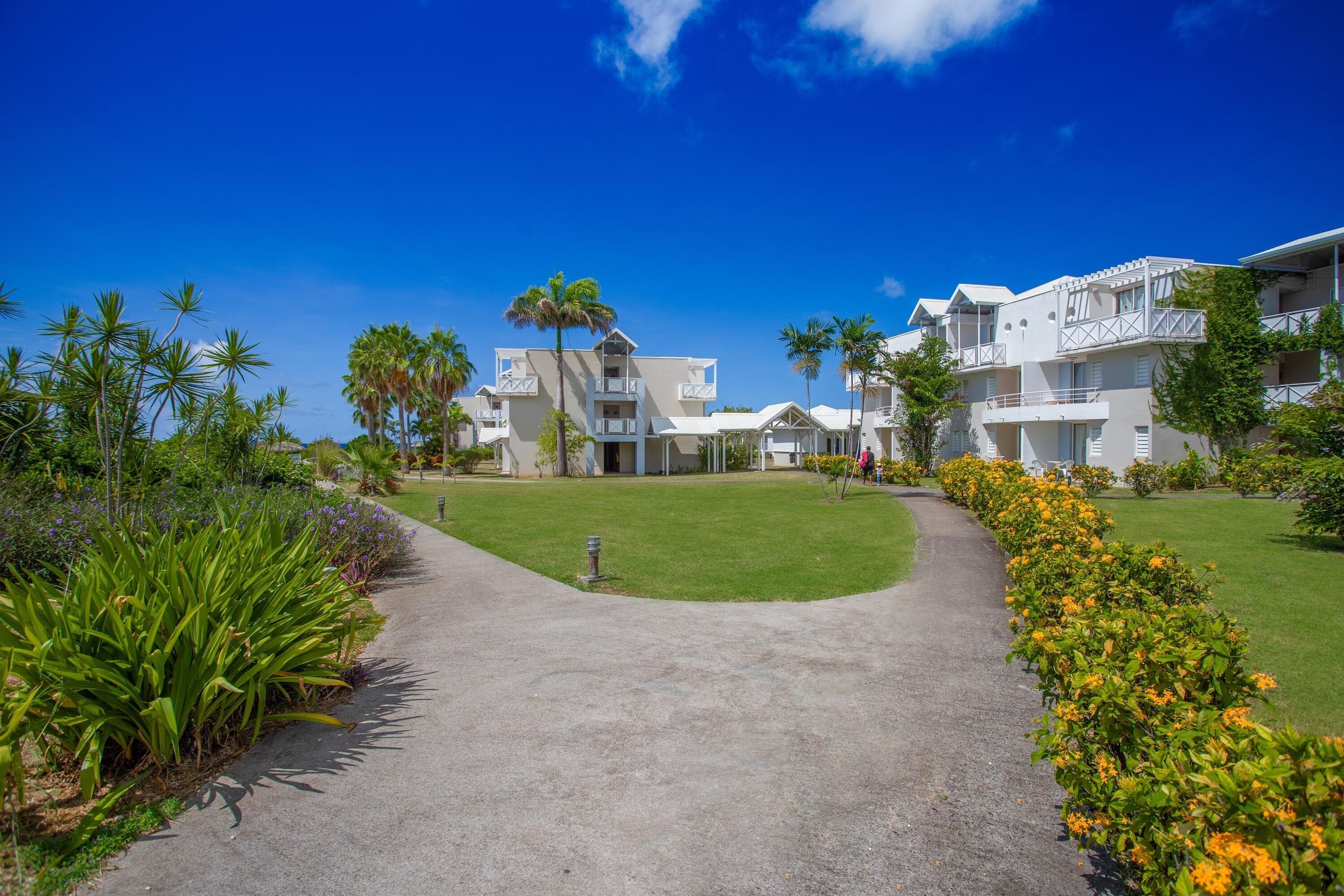A pathway leading to a white building with balconies, surrounded by greenery and palm trees.